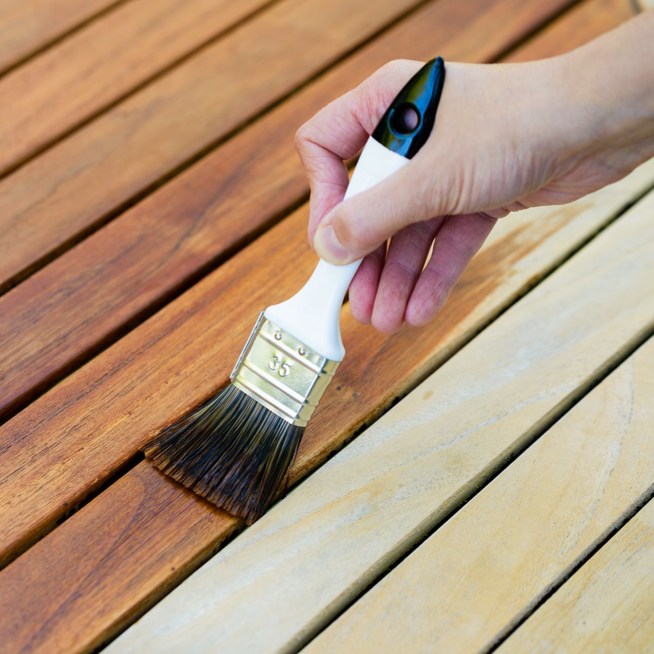 Une main utilise un pinceau pour appliquer une teinture brune riche sur des planches de bois brut, créant ainsi un contraste avec le bois brut.