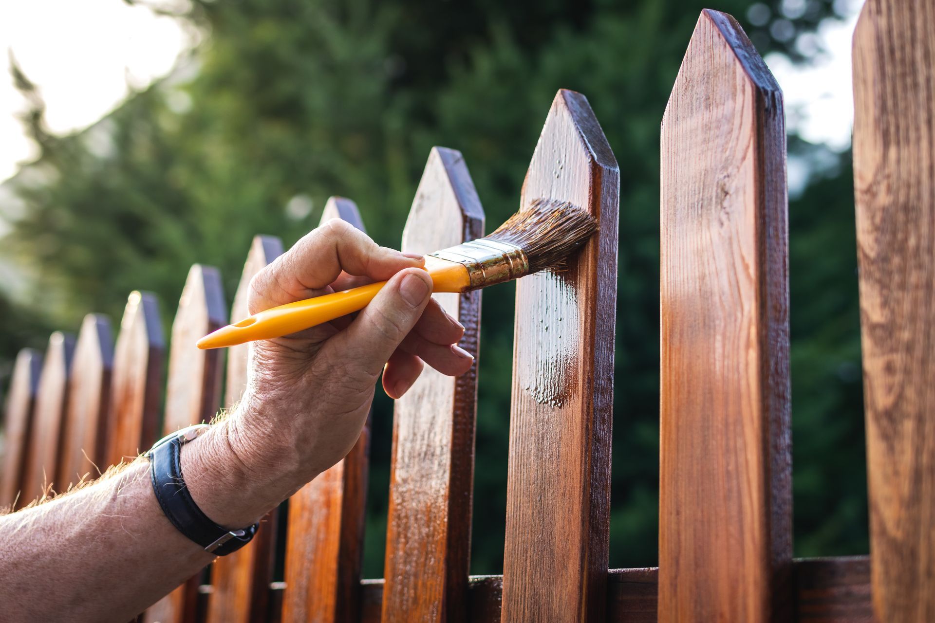 Une main utilise un pinceau à manche jaune pour appliquer un vernis transparent sur le dessus d'un poteau de clôture en bois.