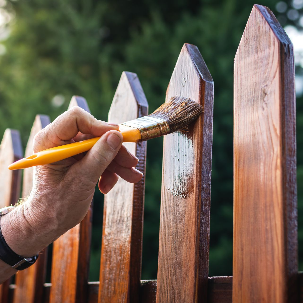 Une main utilise un pinceau à manche jaune pour appliquer une teinture transparente sur une clôture en bois à piquets, en extérieur.