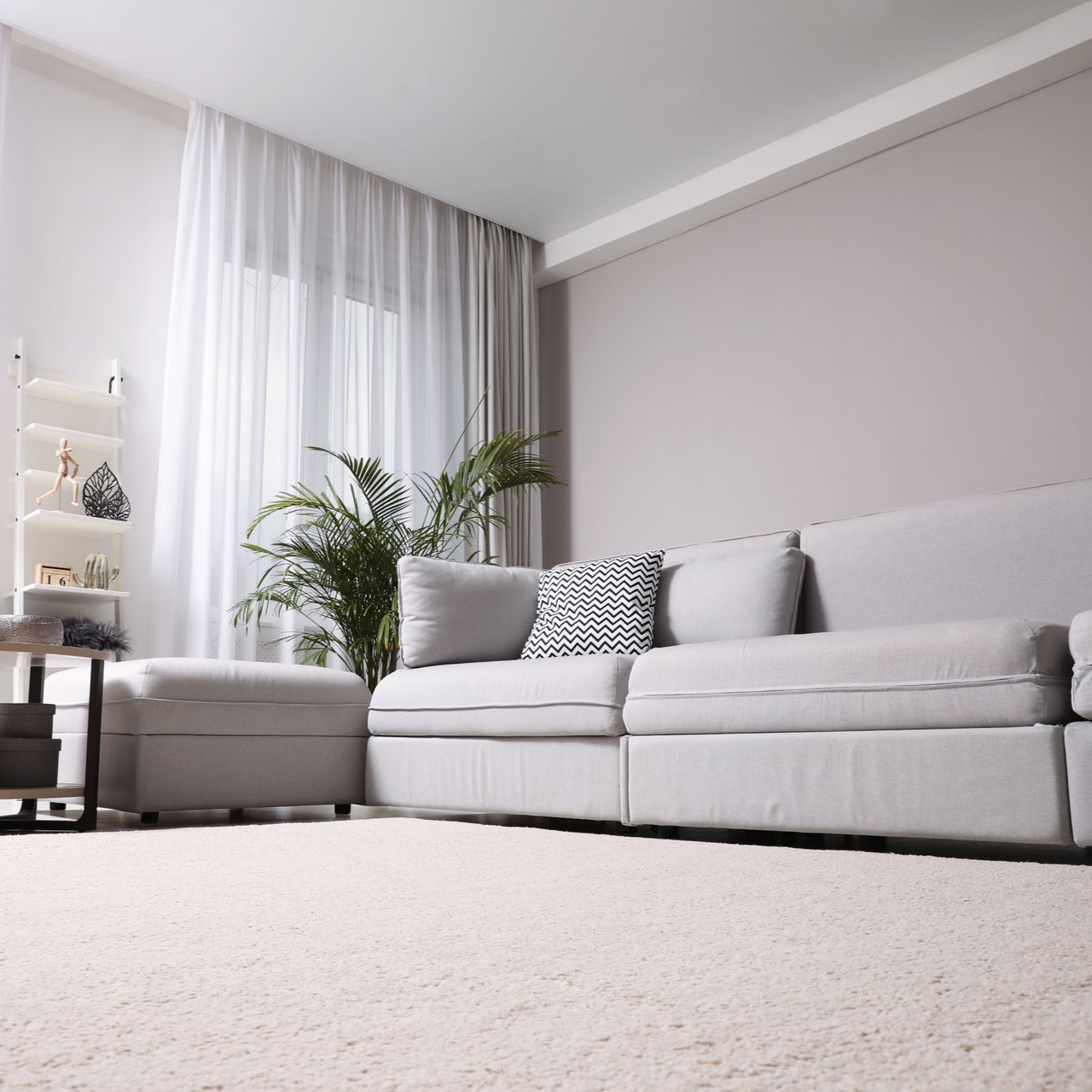 Un salon moderne et minimaliste avec un canapé gris clair, un coussin à motifs, une plante verte et des rideaux blancs.