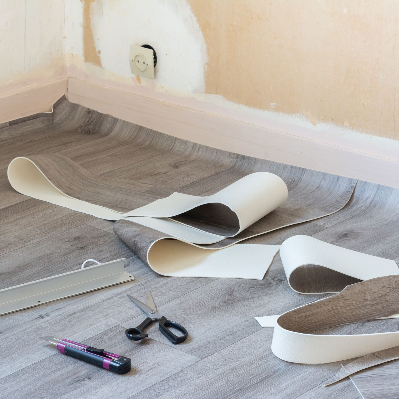 Un rouleau de revêtement de sol en vinyle gris imitation bois est installé dans un coin, avec un cutter et des ciseaux posés au sol.
