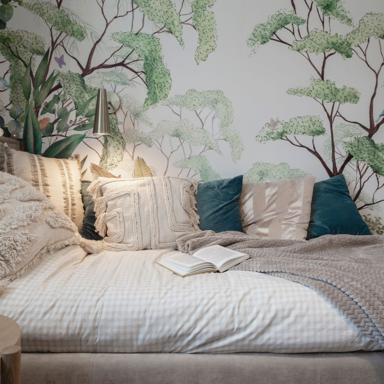 Un lit douillet avec une couette à carreaux vichy, des oreillers texturés et une couverture en grosse maille, le tout adossé à une fresque murale à motifs botaniques.