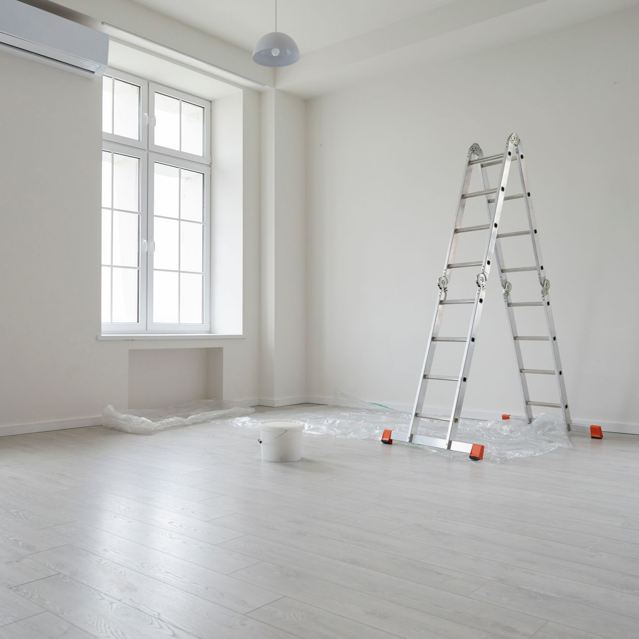 Une pièce lumineuse aux murs blancs, une grande fenêtre, un parquet clair, un escabeau et un seau de peinture posé sur une bâche.