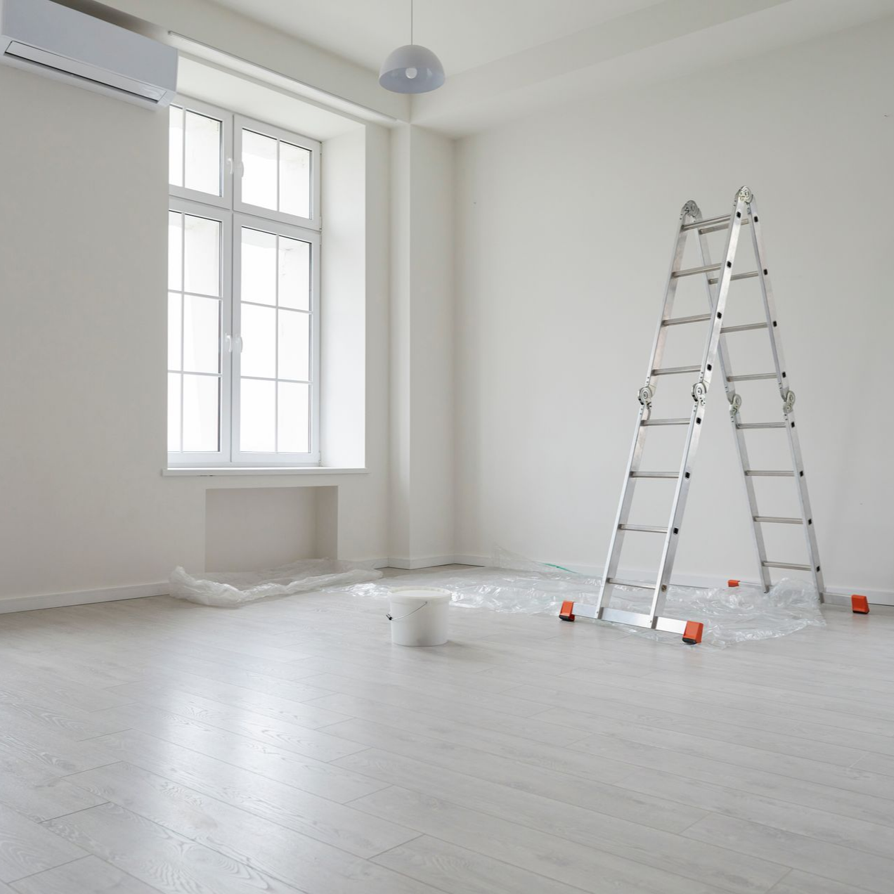 Une échelle métallique se dresse dans une pièce vide aux murs blancs, avec une fenêtre, un luminaire et une bâche en plastique au sol.