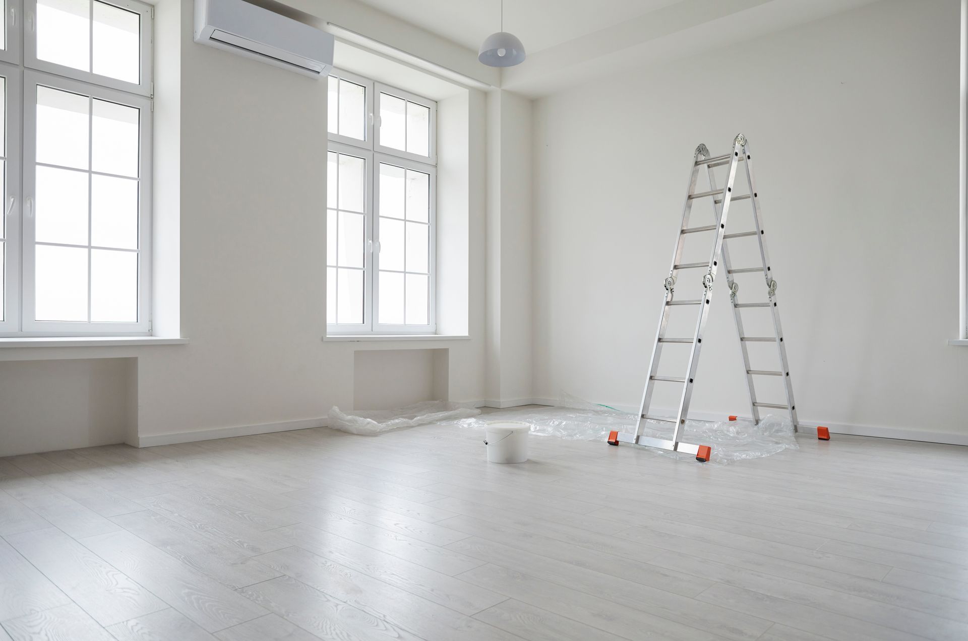 Une pièce vide aux murs blancs, avec de grandes fenêtres, un escabeau en métal et un seau de peinture posé sur une bâche.