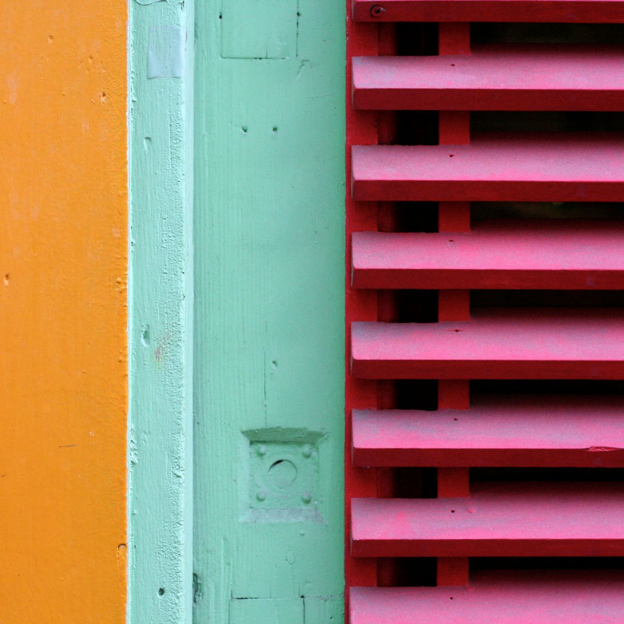 Un mur orange texturé jouxte une moulure verticale vert menthe et des volets horizontaux rouges.