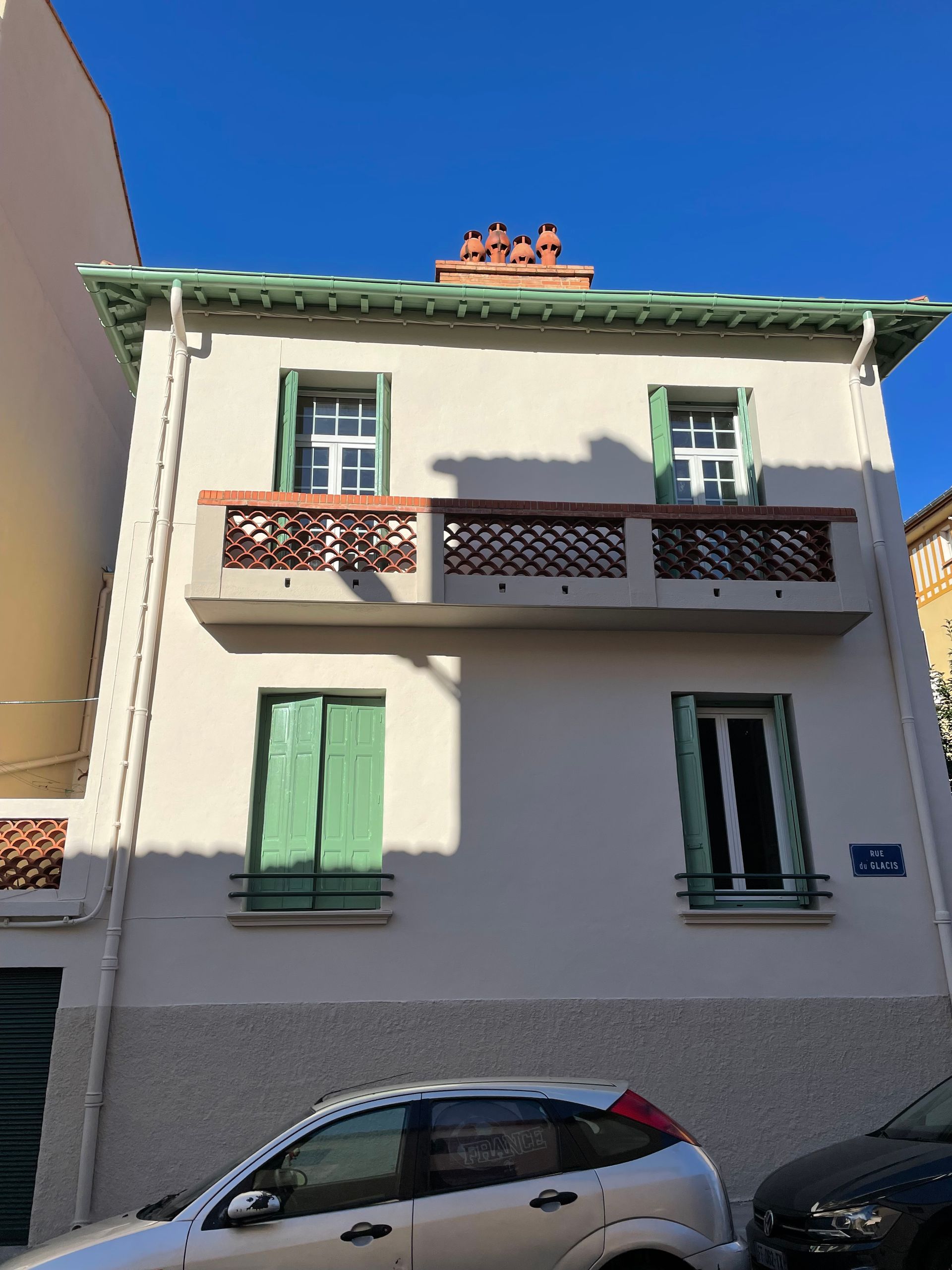 Une maison claire de deux étages avec des volets verts, un balcon et un toit de tuiles, sous un ciel bleu azur.