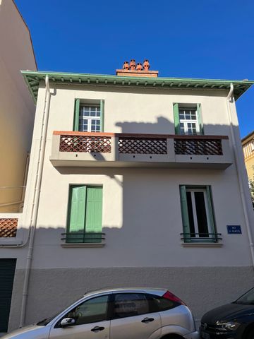 Une maison en stuc de deux étages avec des volets verts et un balcon marron, vue depuis une rue avec des voitures garées sur fond de ciel bleu.