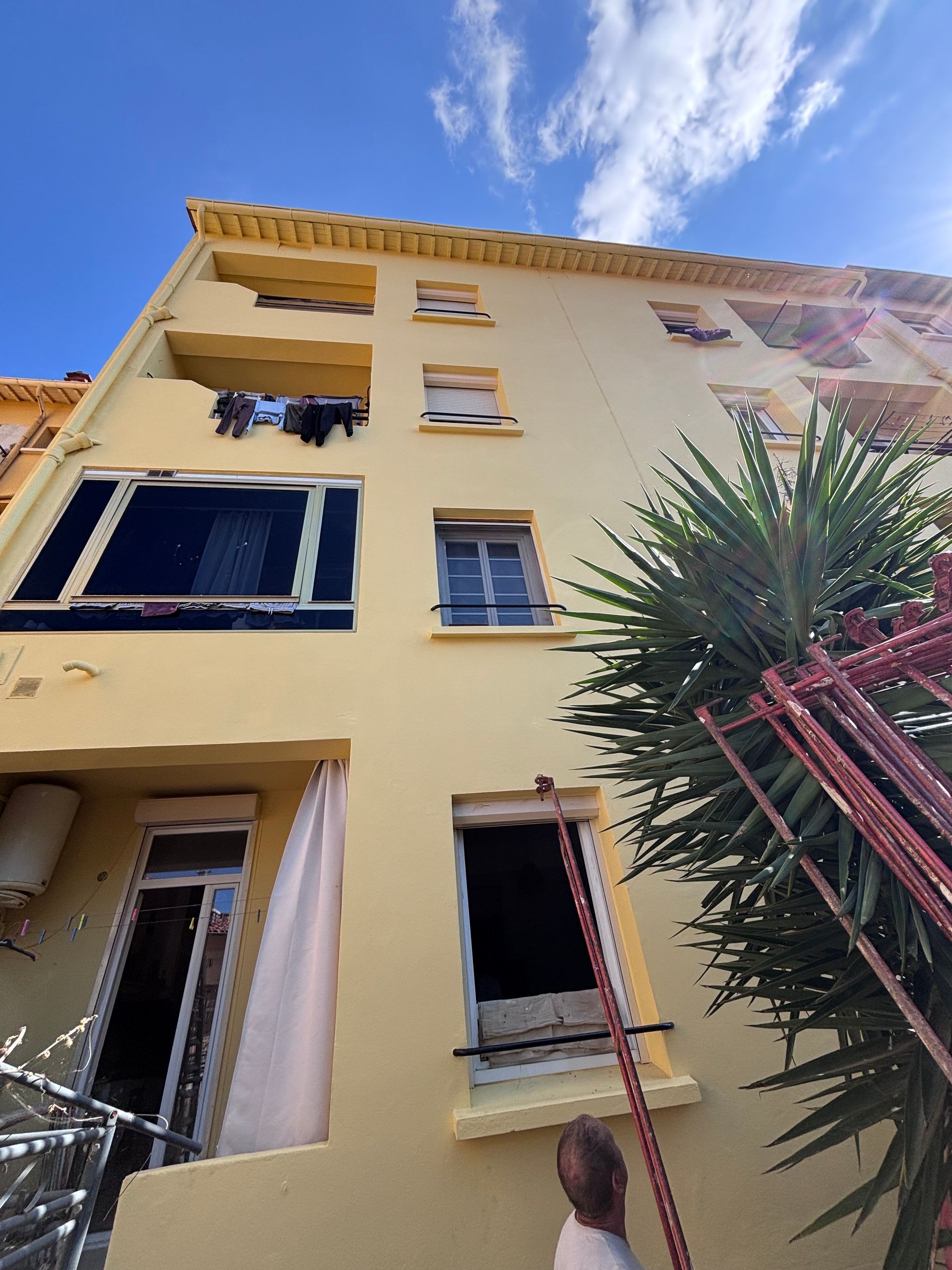 Vue en contreplongée de la façade d'un immeuble d'appartements jaune avec des balcons, une fenêtre sombre et une échelle appuyée contre un mur.