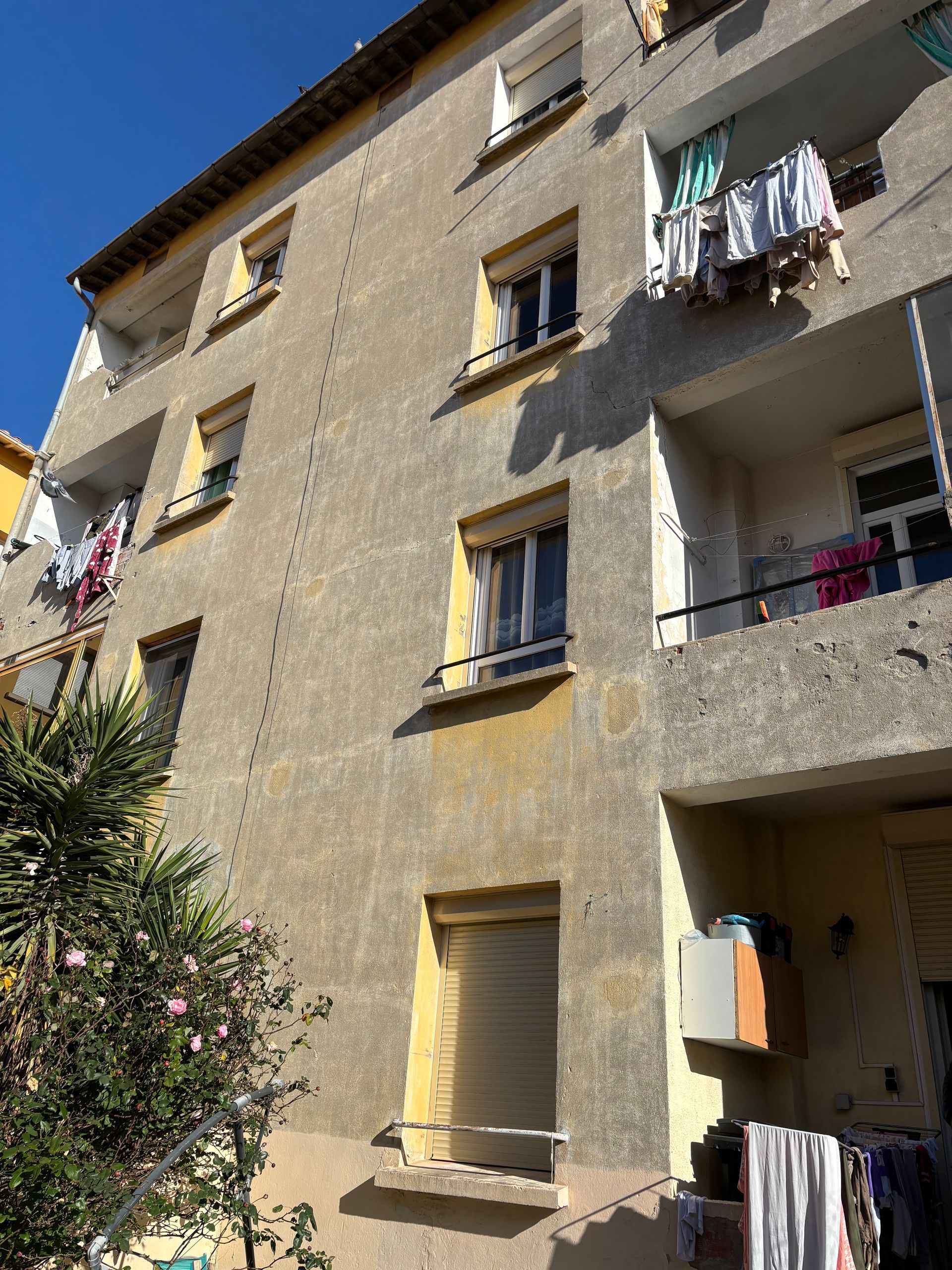 Un immeuble d'appartements à plusieurs étages avec des murs, des fenêtres et des balcons à la texture beige, où du linge sèche au soleil.