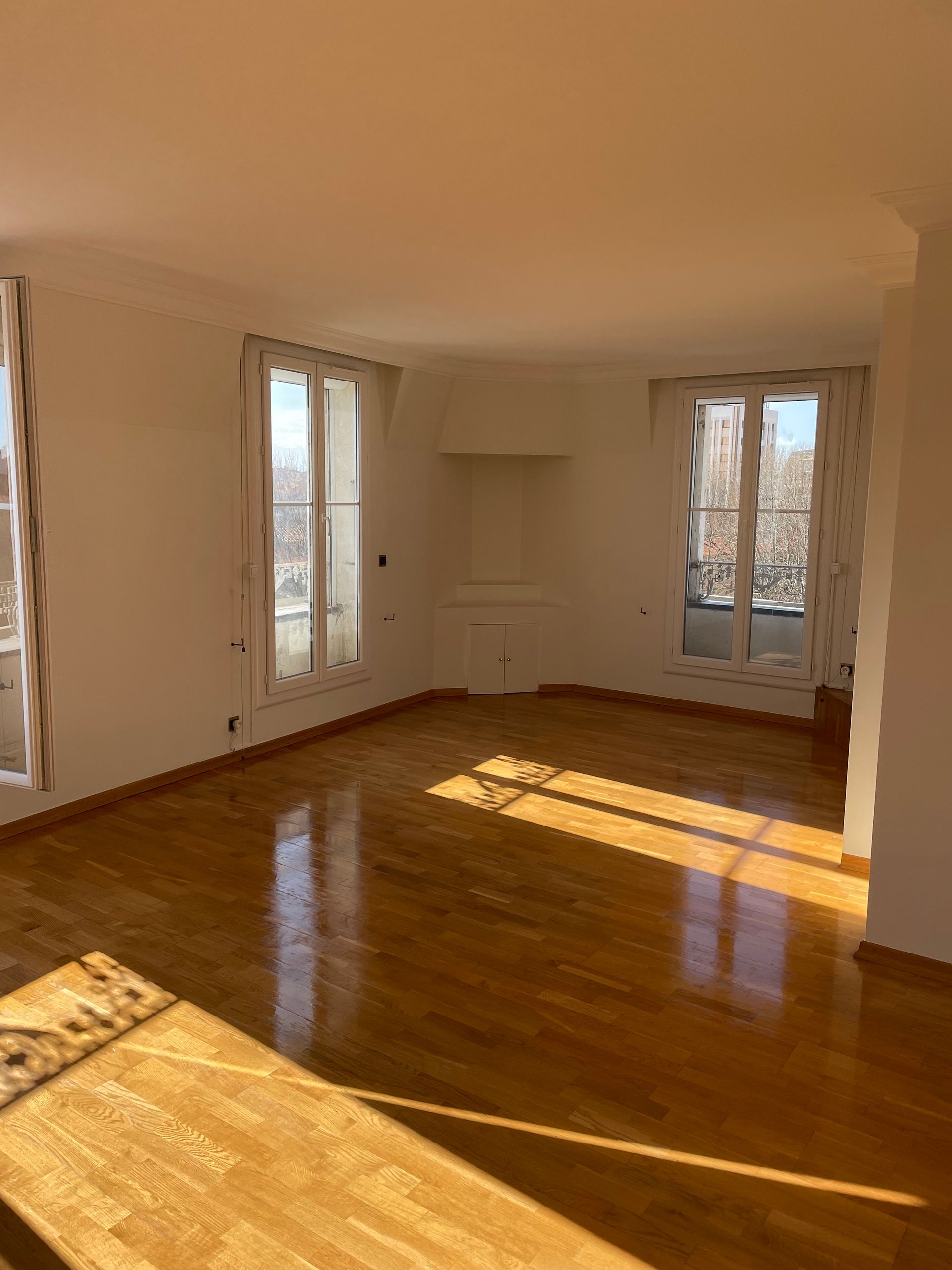 Une pièce vide et lumineuse, avec un parquet ciré, des murs blancs et deux grandes fenêtres laissant entrer la lumière du soleil.
