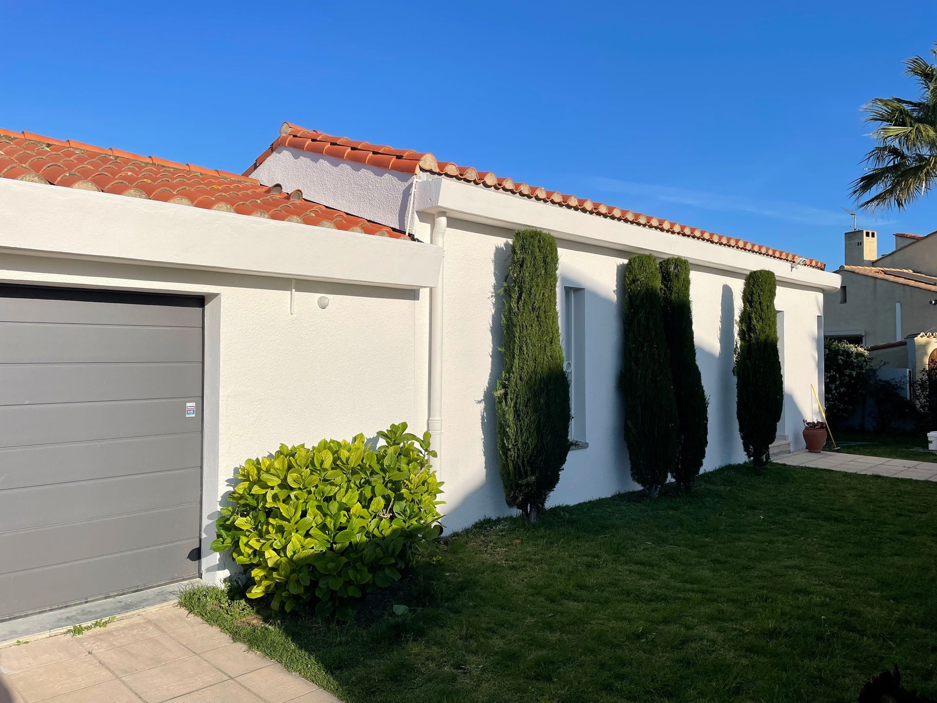 Une maison blanche avec une porte de garage grise, un toit de tuiles rouges et trois grands cyprès verts dans la cour avant.