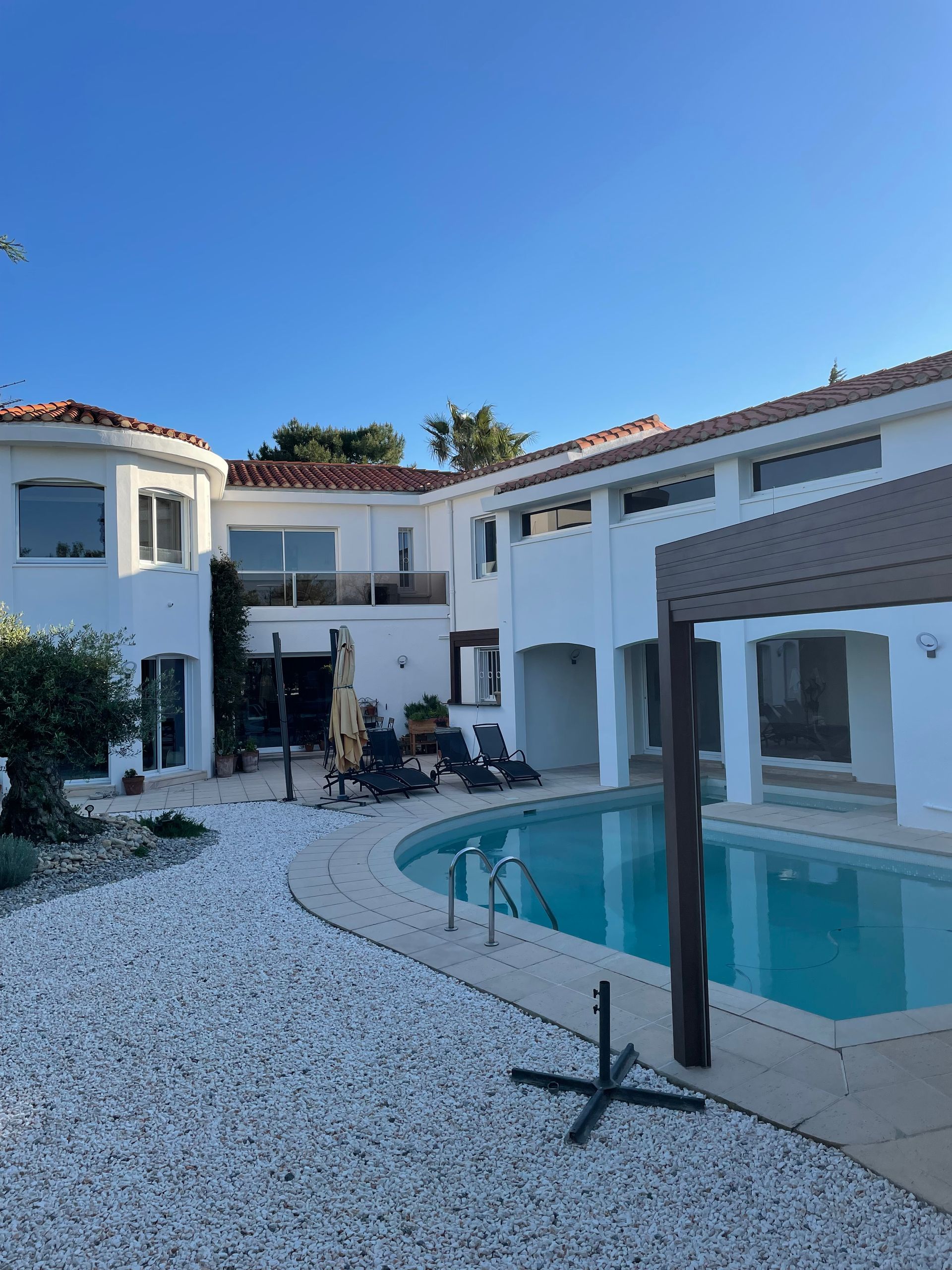 Une maison blanche à plusieurs étages avec une piscine bleue et une terrasse en gravier blanc sous un ciel d'un bleu limpide.