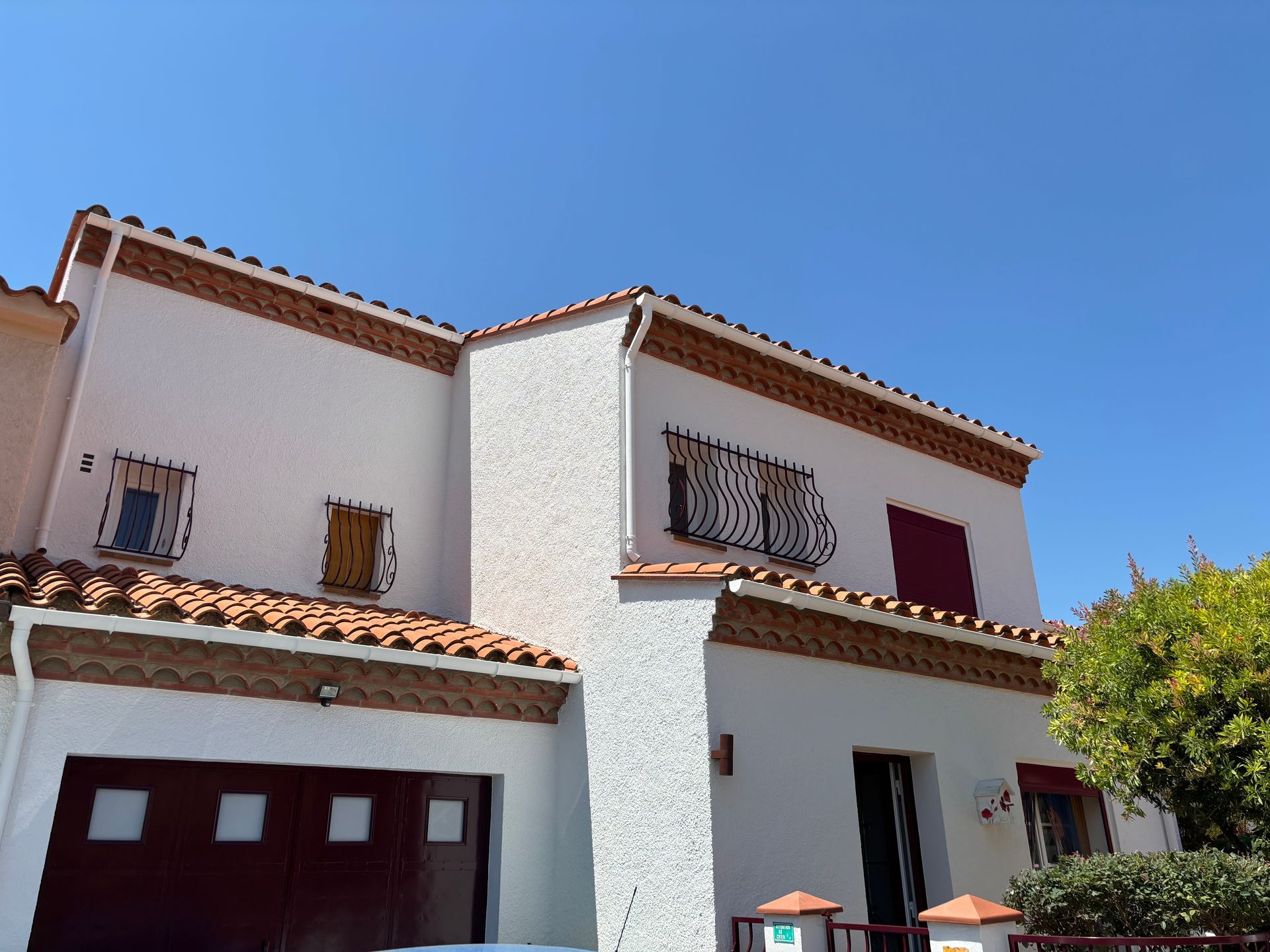 Une maison blanche à deux étages avec une façade texturée, des toits de tuiles en terre cuite, des volets sombres et un garage sous un ciel dégagé.