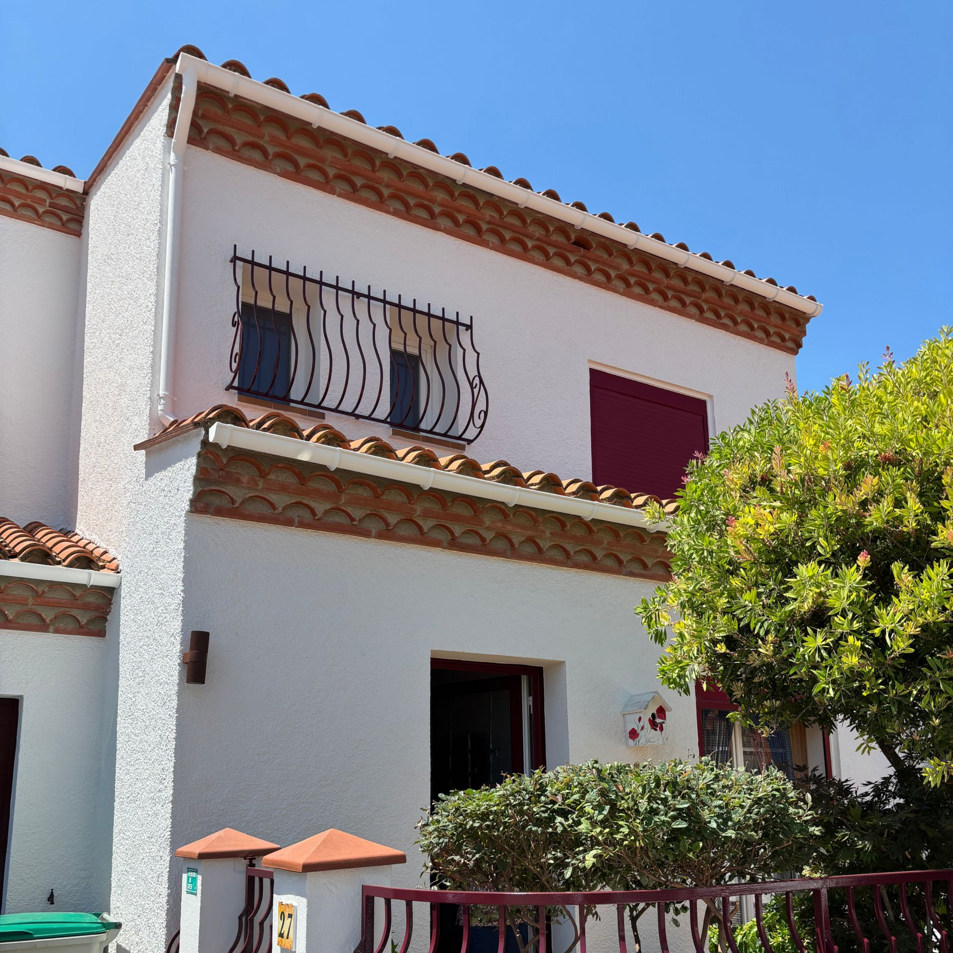 Une maison blanche à deux étages avec un toit de tuiles en terre cuite, une grille de fenêtre noire et un volet rouge, sous un ciel bleu limpide.