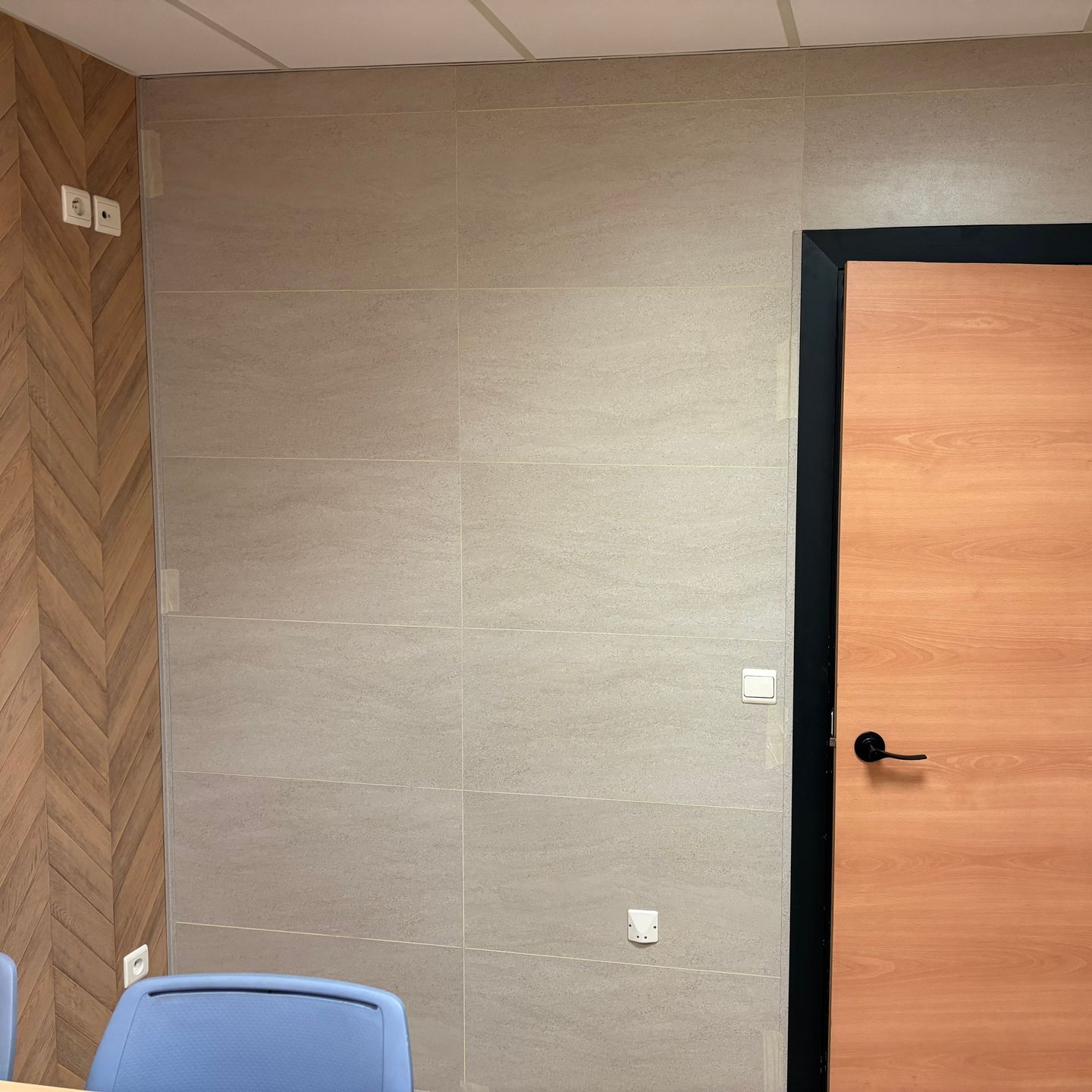 Un mur carrelé de gris jouxte une porte en bois clair avec un cadre et une poignée noirs, et une chaise bleue au premier plan.