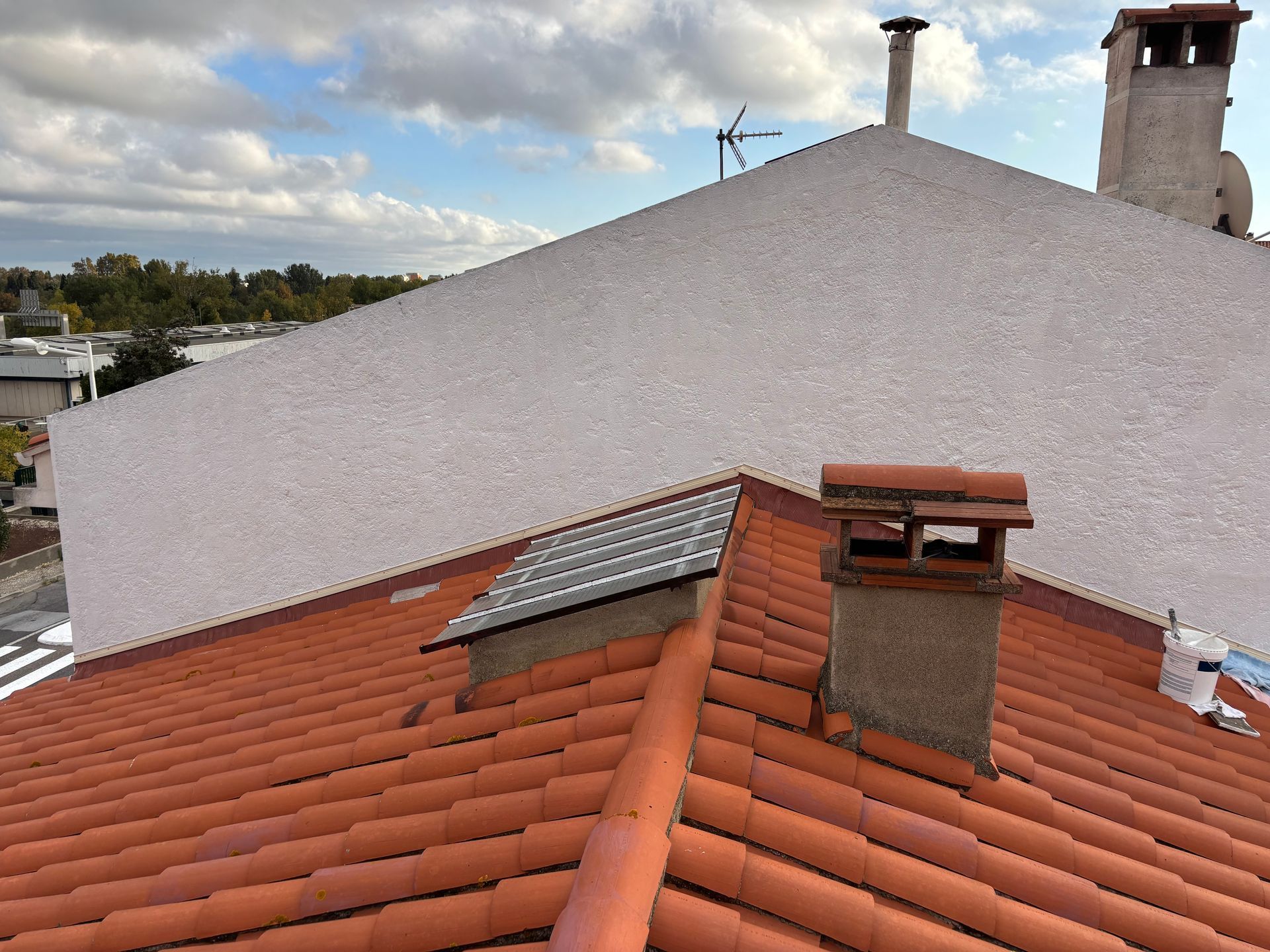 Vue d'un toit de tuiles brun rougeâtre avec une cheminée et une petite lucarne, se détachant sur un grand mur extérieur blanc.