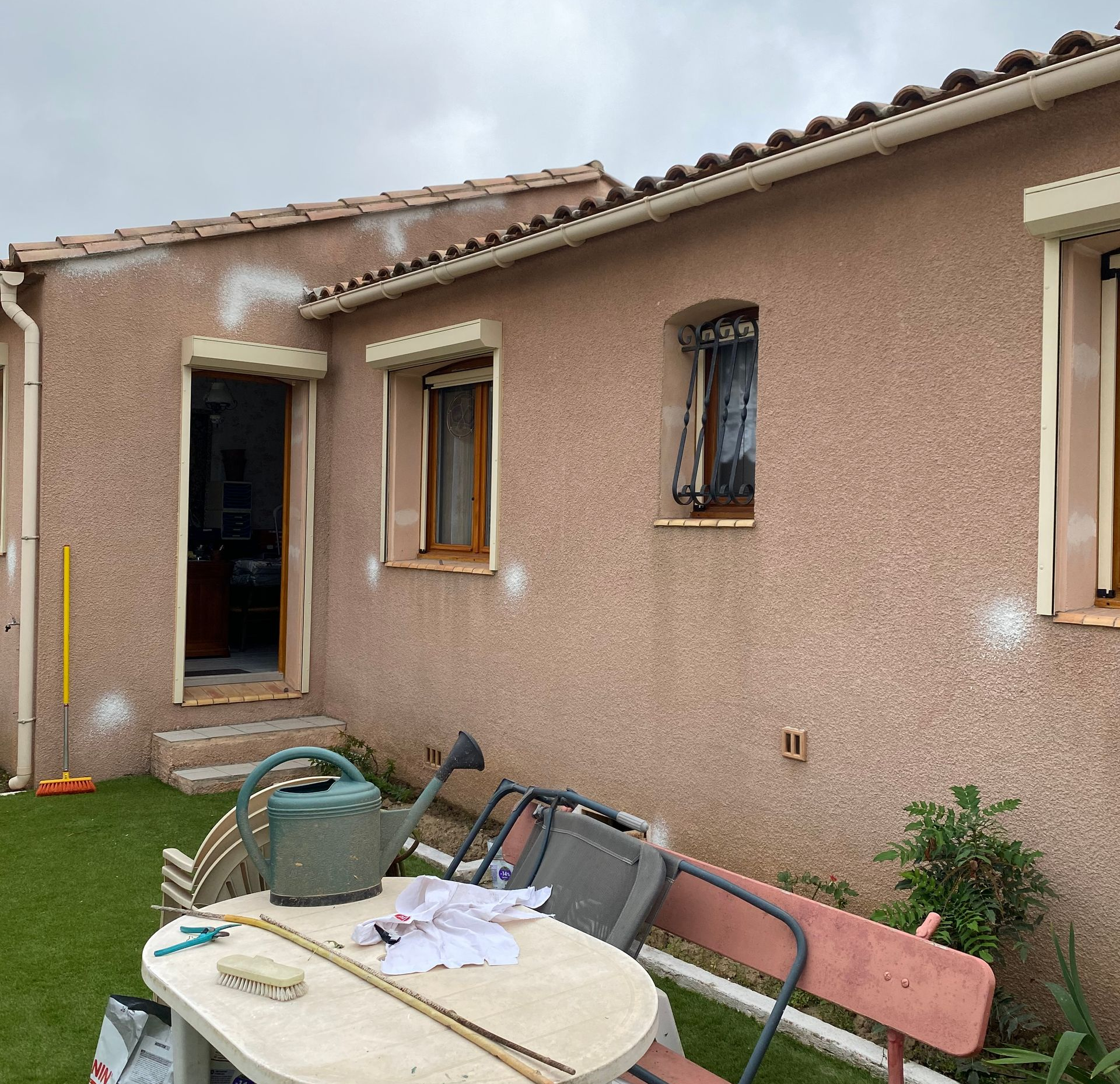 L'extérieur d'une maison en stuc beige avec des fenêtres, une porte et une table extérieure blanche avec un arrosoir au premier plan.