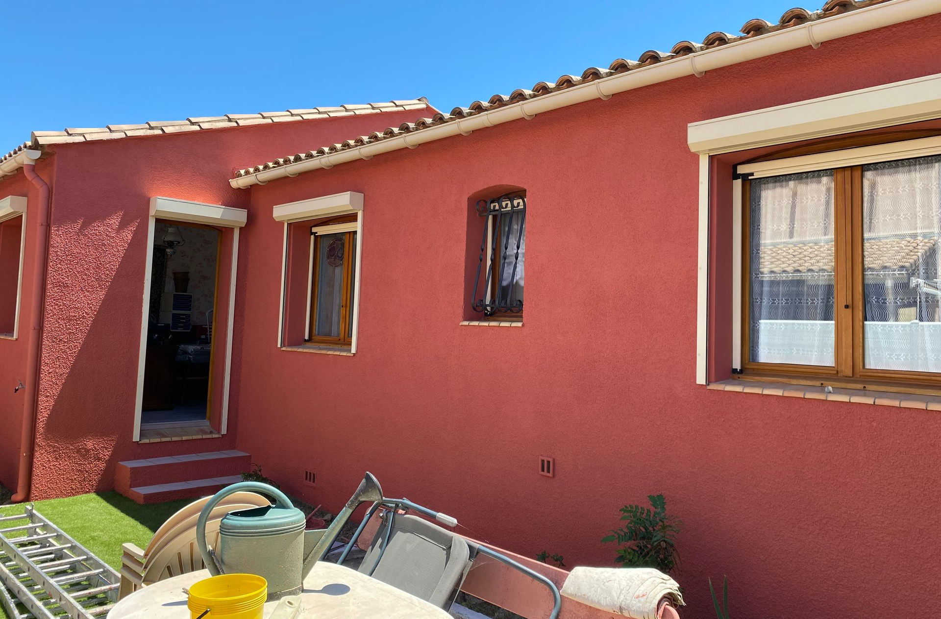 L'extérieur d'une maison couleur terre cuite, avec une porte, plusieurs fenêtres et du mobilier de jardin, par une journée ensoleillée.