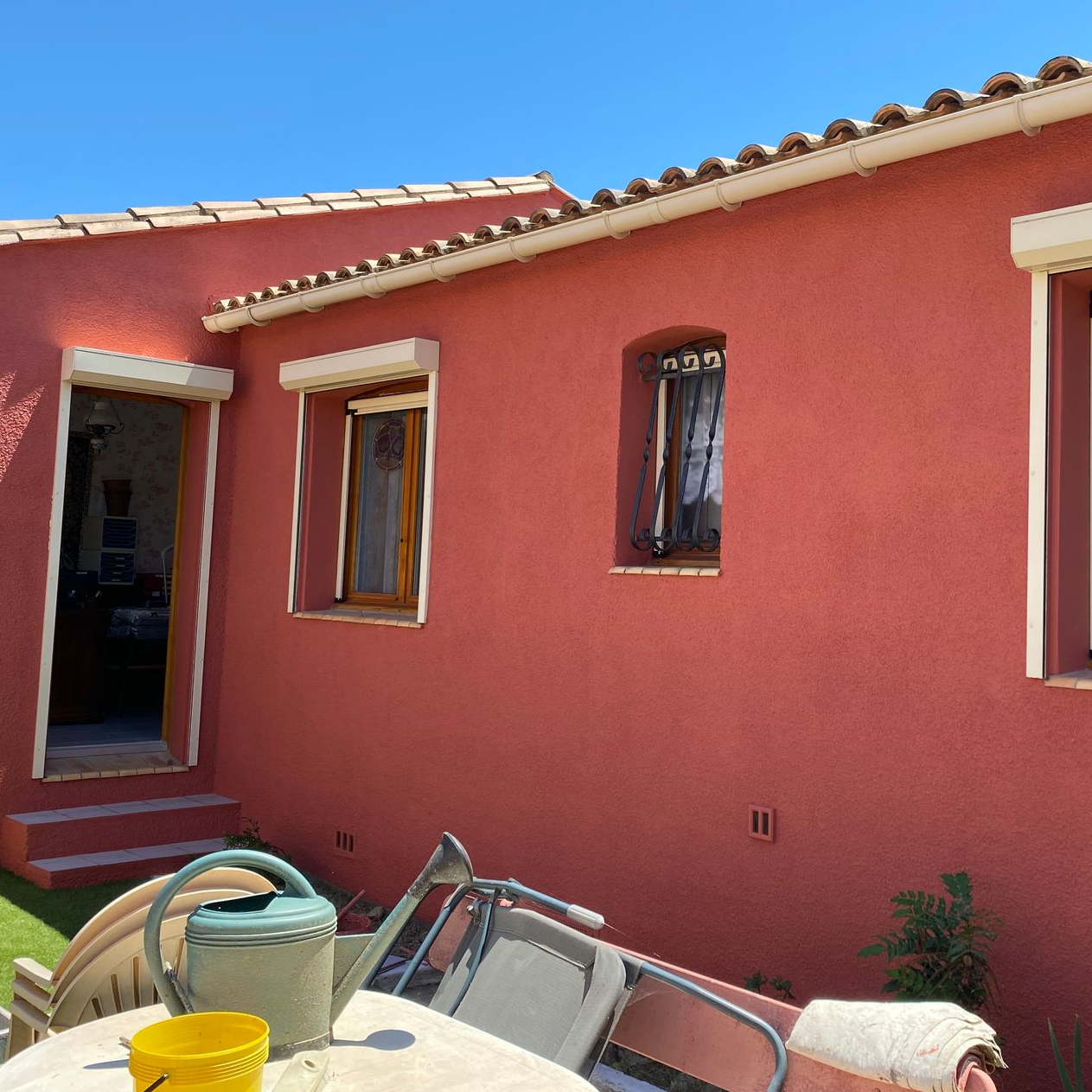 Vue extérieure d'une maison couleur terre cuite avec deux fenêtres et une porte, et du mobilier de jardin sur une terrasse.