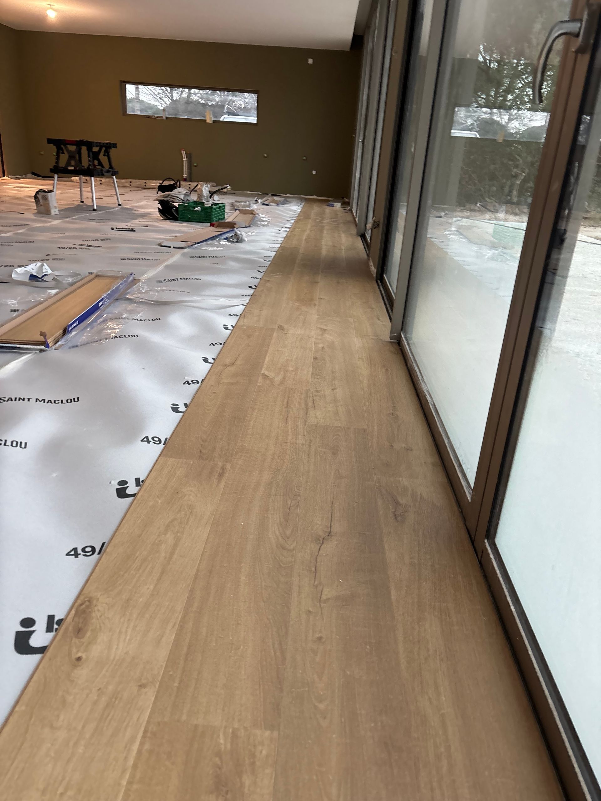 Une personne agenouillée sur un sol sombre installe des lames de parquet stratifié clair, un marteau à proximité.