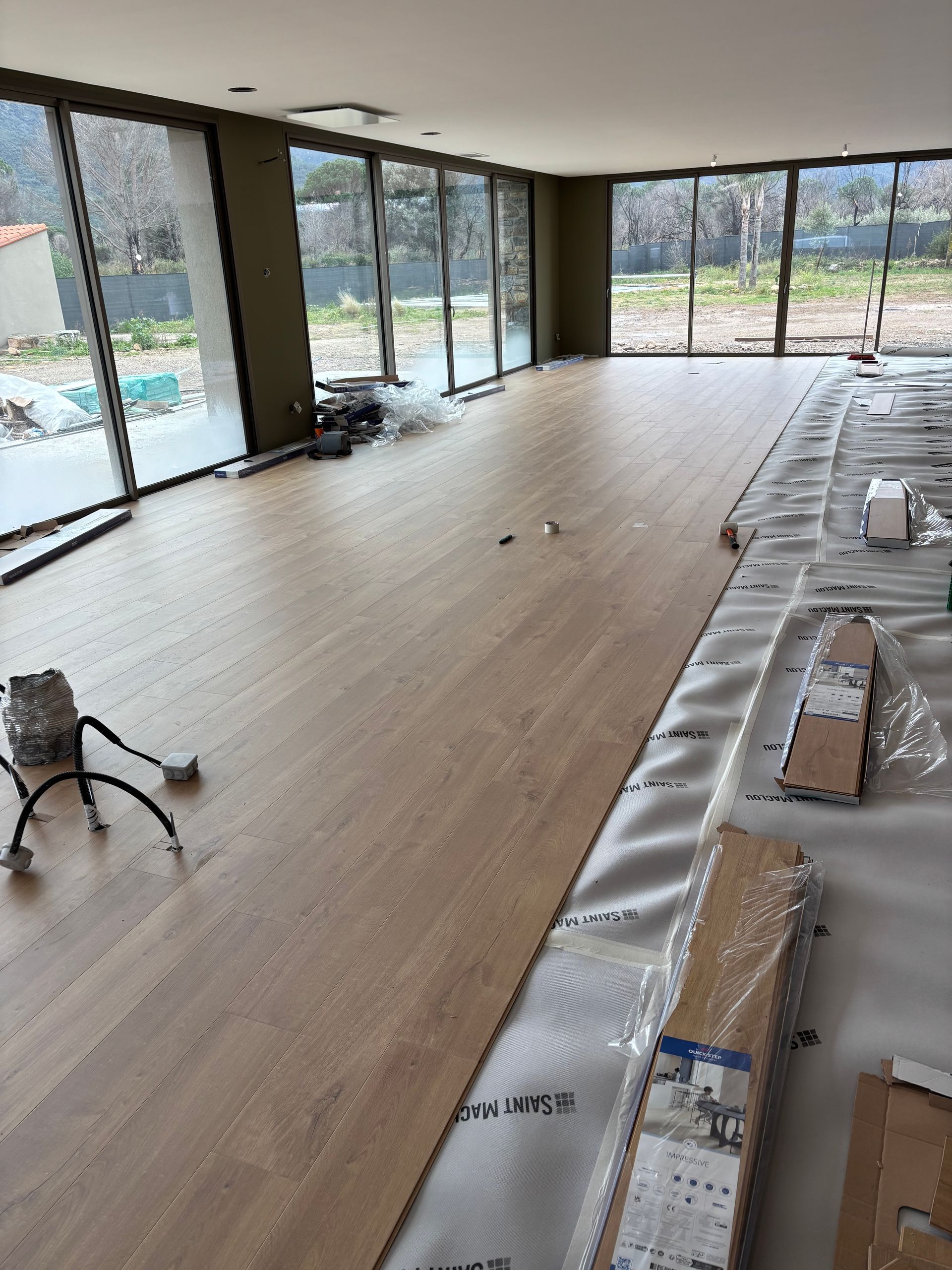 Vue en contreplongée de lames de parquet imitation bois dans une teinte brune chaude, avec un salon flou en arrière-plan.