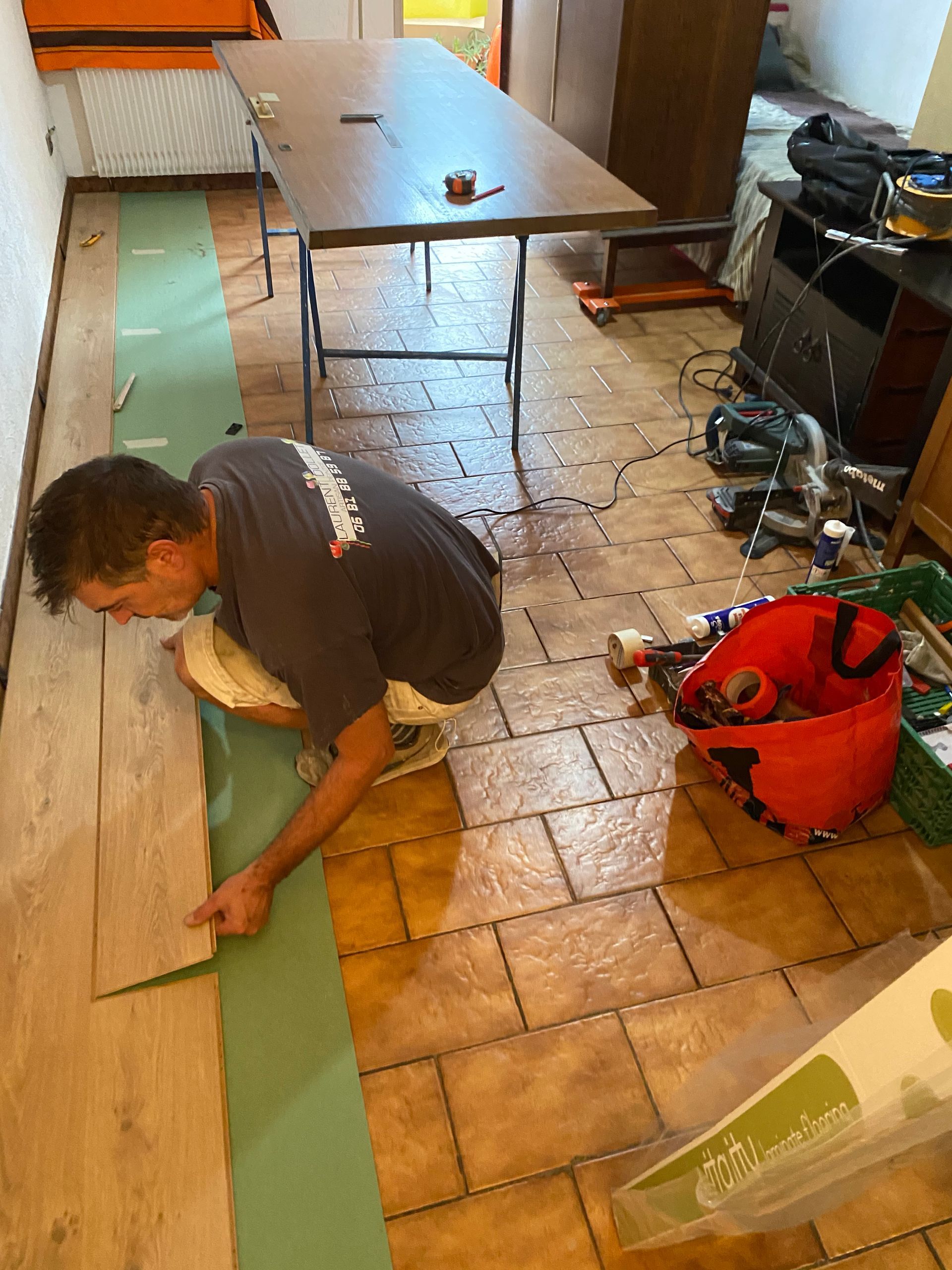Une personne agenouillée sur une sous-couche de sol verte, en train d'installer un revêtement de sol stratifié imitation bois sur un carrelage existant.