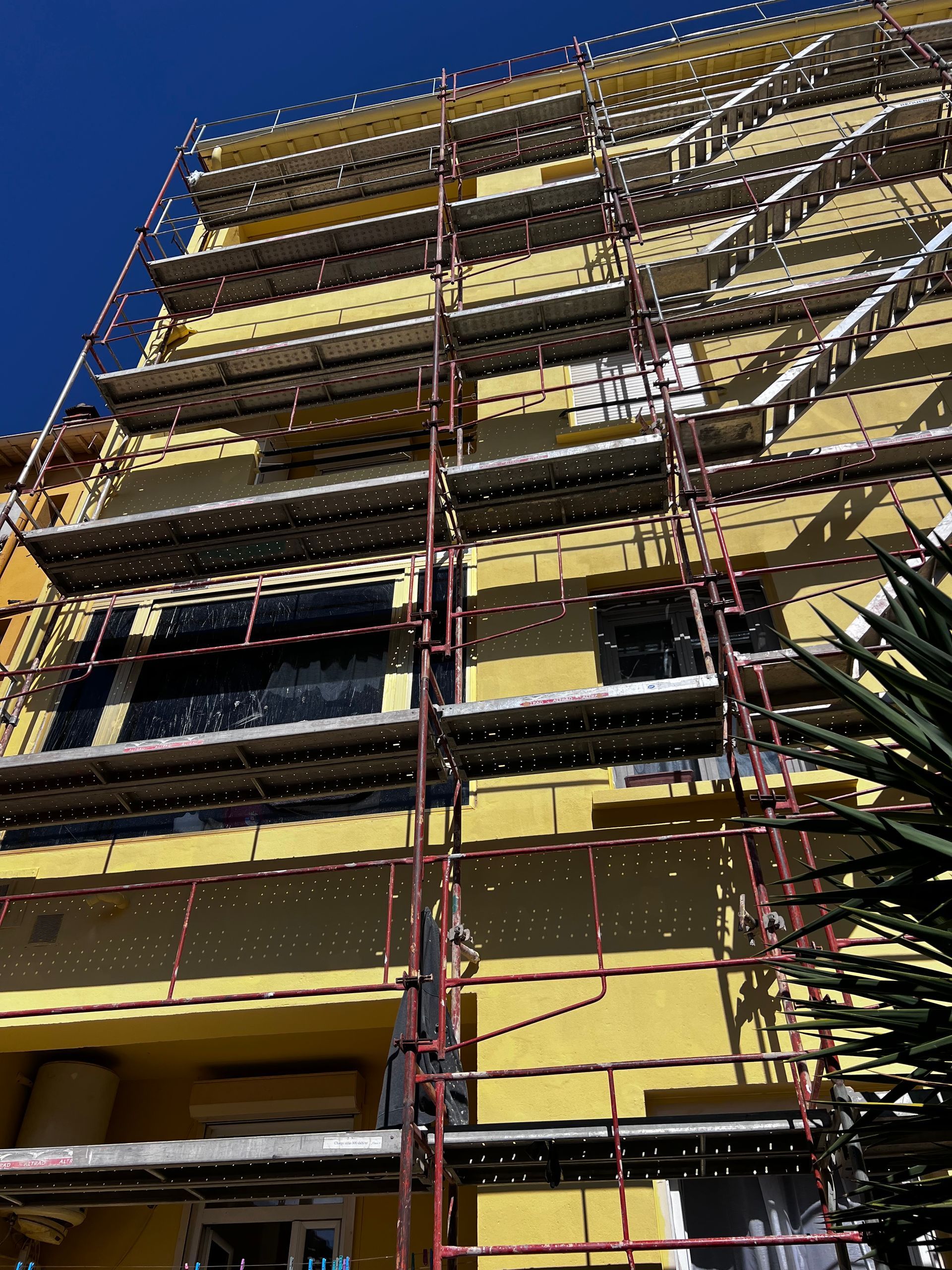 L'extérieur d'un bâtiment jaune est recouvert d'une structure d'échafaudage métallique à plusieurs niveaux, sous un ciel bleu dégagé.