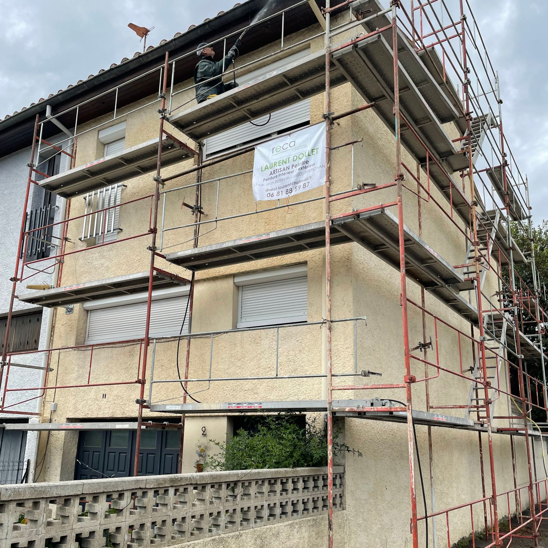 Un immeuble à plusieurs étages, recouvert d'échafaudages pour des rénovations extérieures, avec un ouvrier visible sur la plateforme supérieure.