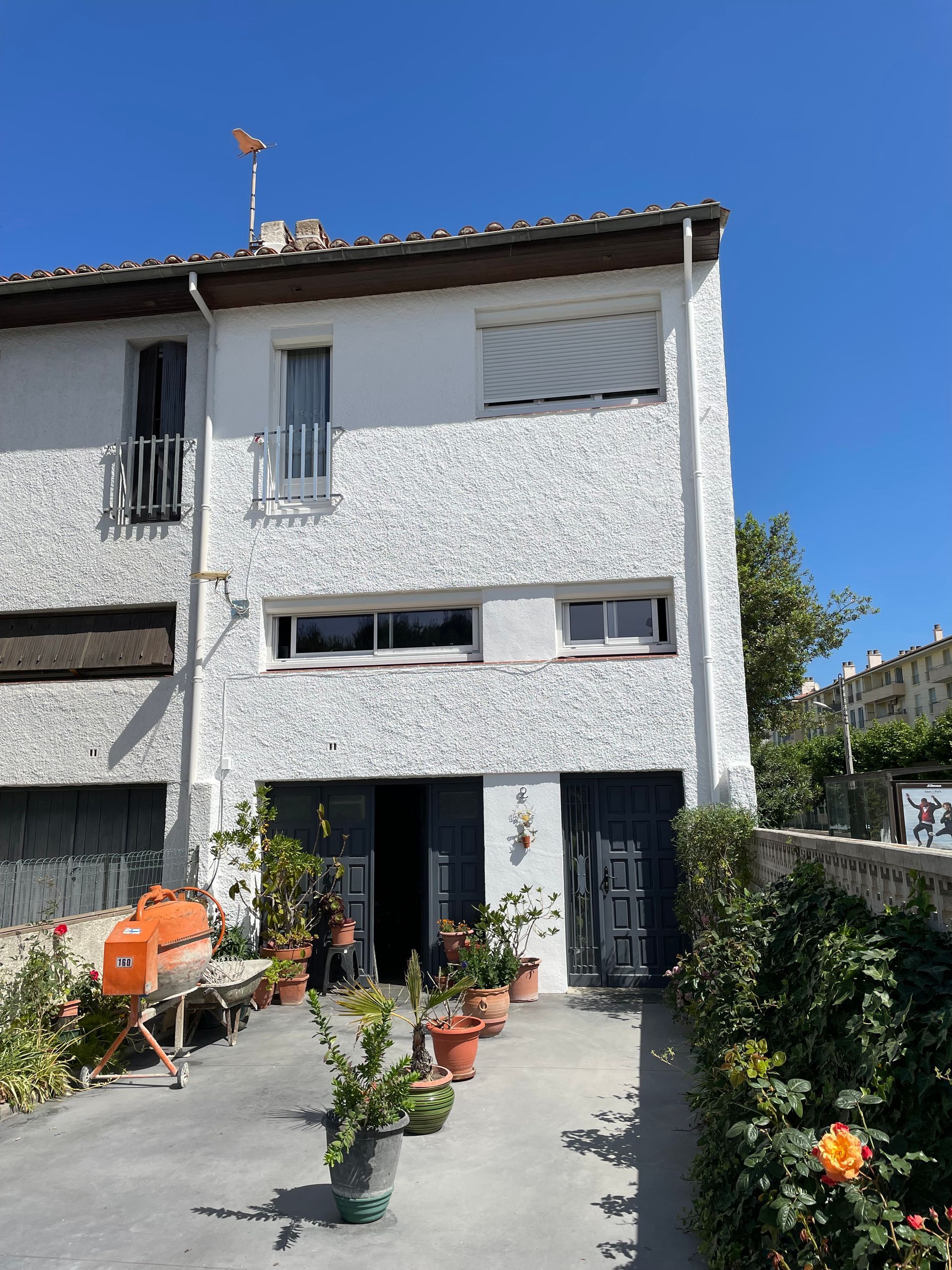 Maison blanche à deux étages avec des murs texturés, un patio gris avec des plantes en pot et un barbecue orange sous un ciel bleu.