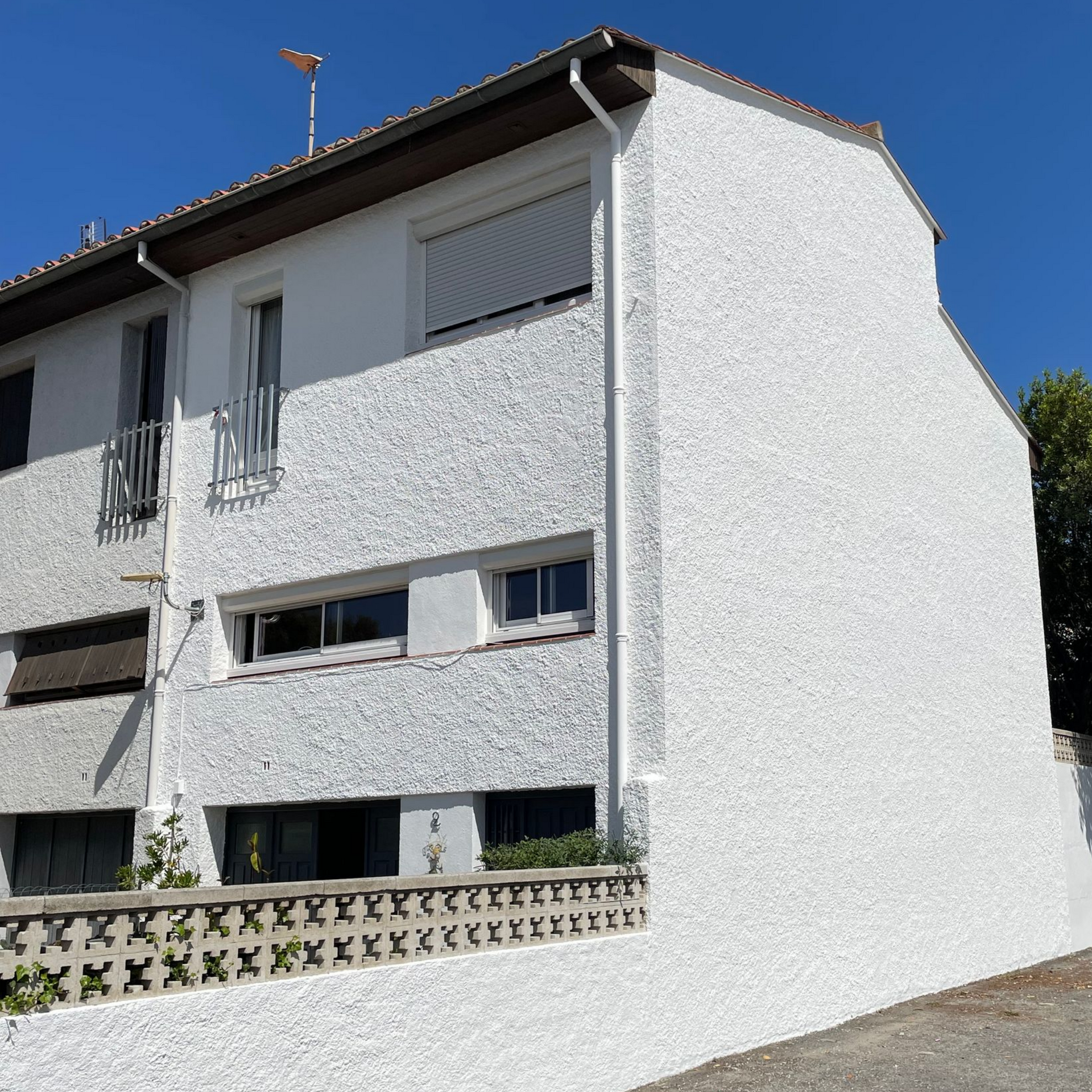 Une maison blanche à deux étages avec une façade texturée, plusieurs fenêtres rectangulaires aux volets fermés et un muret en pierre.