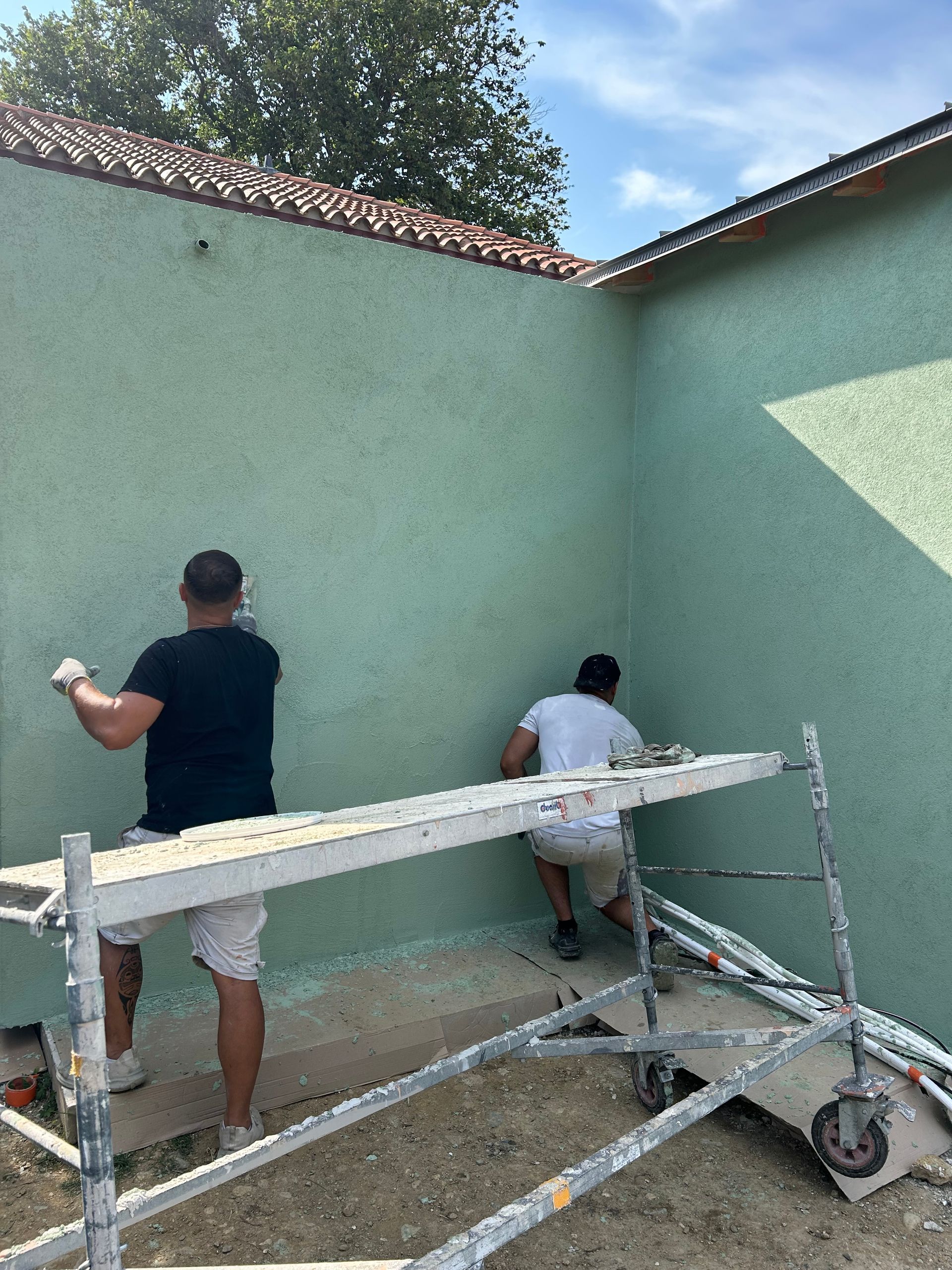 Deux personnes utilisent des outils pour appliquer du stuc sur le mur extérieur vert clair d'un bâtiment, tout en se tenant sur un échafaudage.