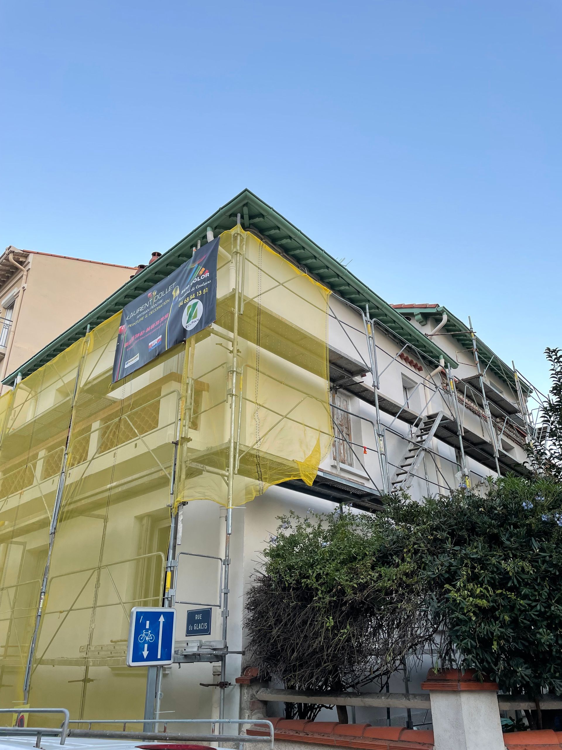 Un immeuble à plusieurs étages, enveloppé d'échafaudages et de filets de chantier jaunes, sous un ciel bleu dégagé.
