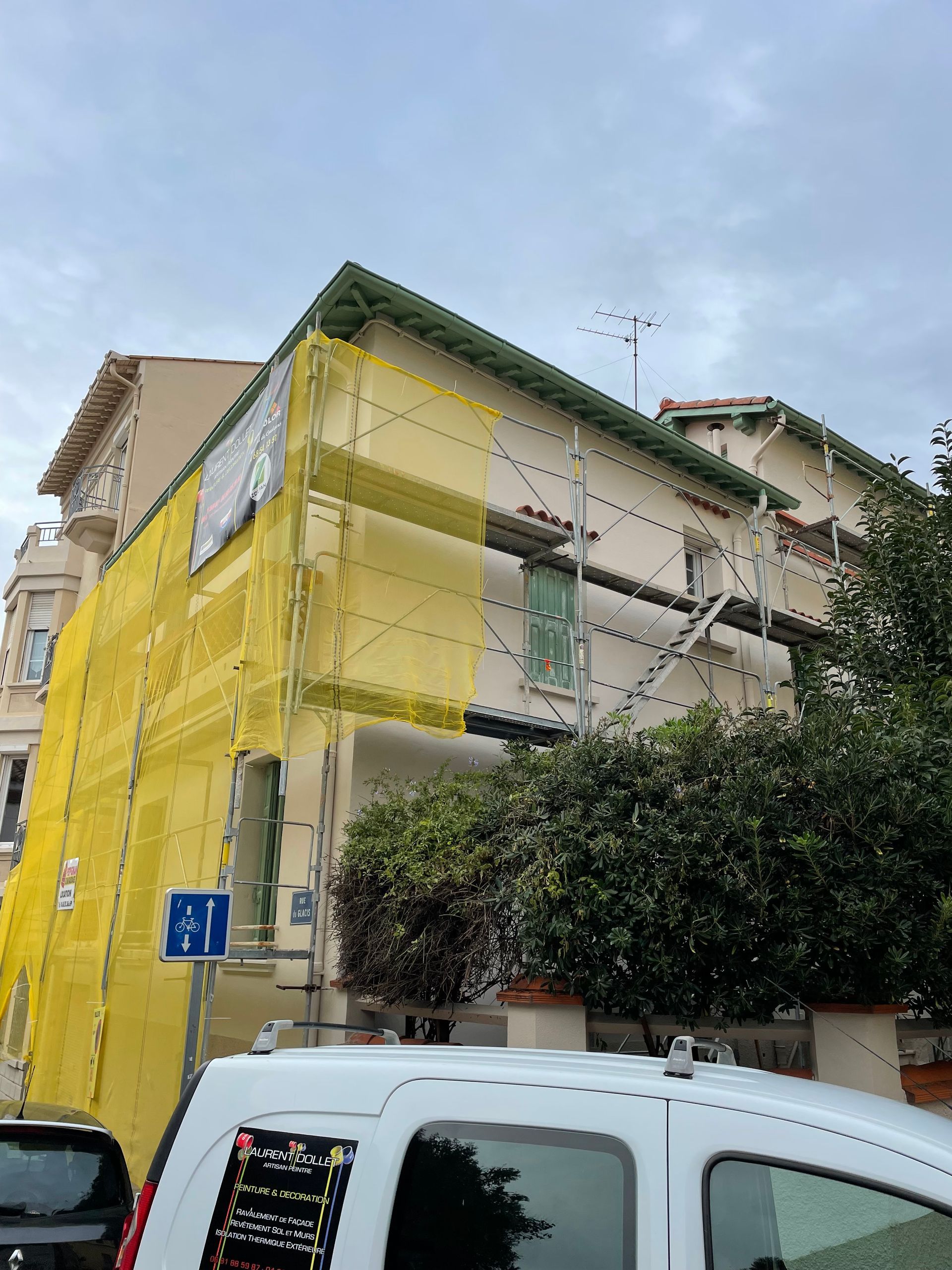 Un immeuble de plusieurs étages en construction, recouvert de filets d'échafaudage jaunes, visible derrière une camionnette de service blanche.