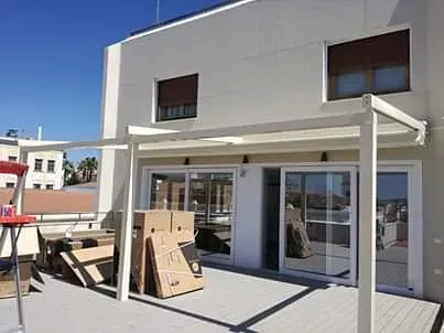 Una pérgola blanca se encuentra sobre una terraza de madera frente a una casa.