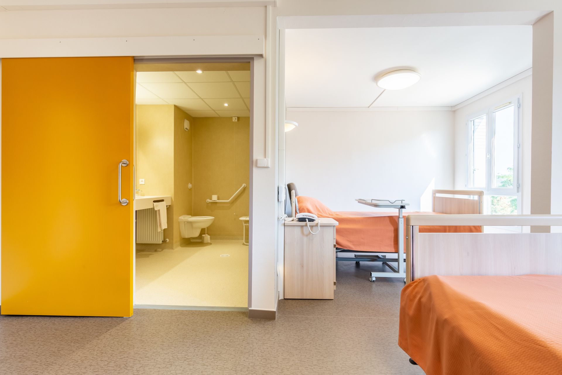 Chambre d’hôpital moderne avec porte coulissante orange, salle de bains, lit, table de chevet et grandes fenêtres lumineuses