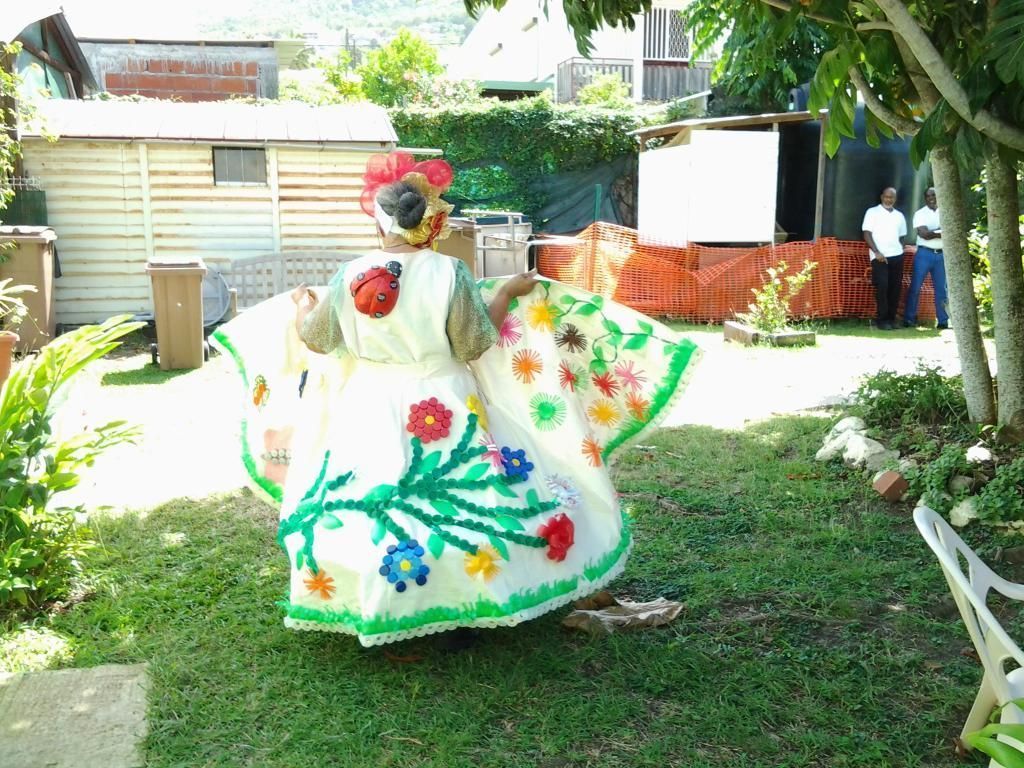 Personne en robe fleurie et colorée, posant dans un jardin herbeux près d’une maison et d’arbres.