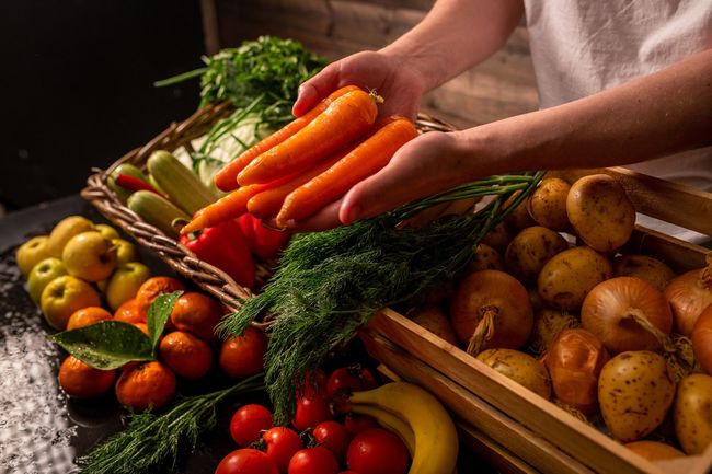 Una persona sostiene un manojo de zanahorias frente a una mesa llena de frutas y verduras.