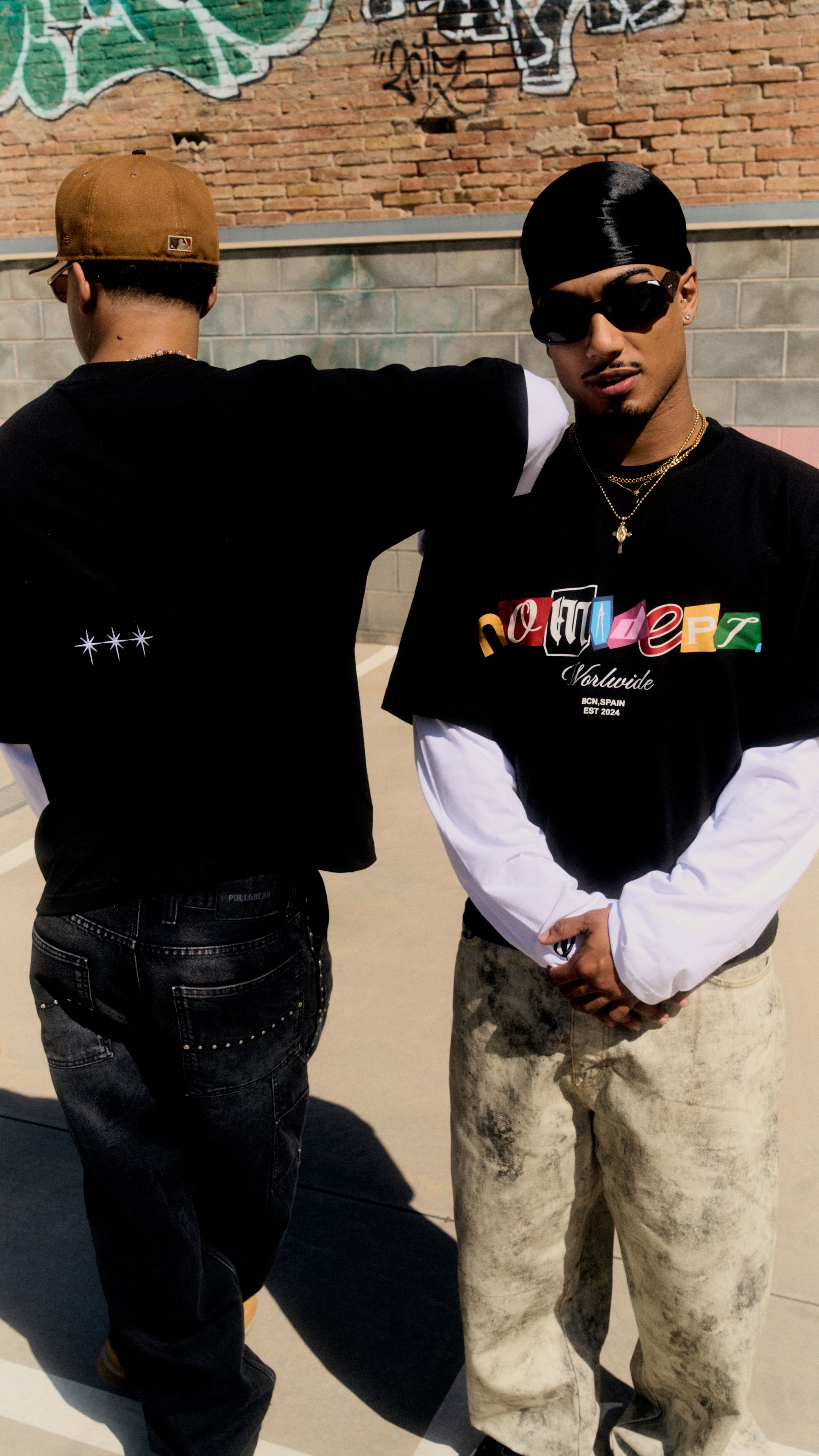 Dos personas posando al aire libre; una lleva un durag y gafas de sol, la otra una gorra de béisbol. Ambas visten