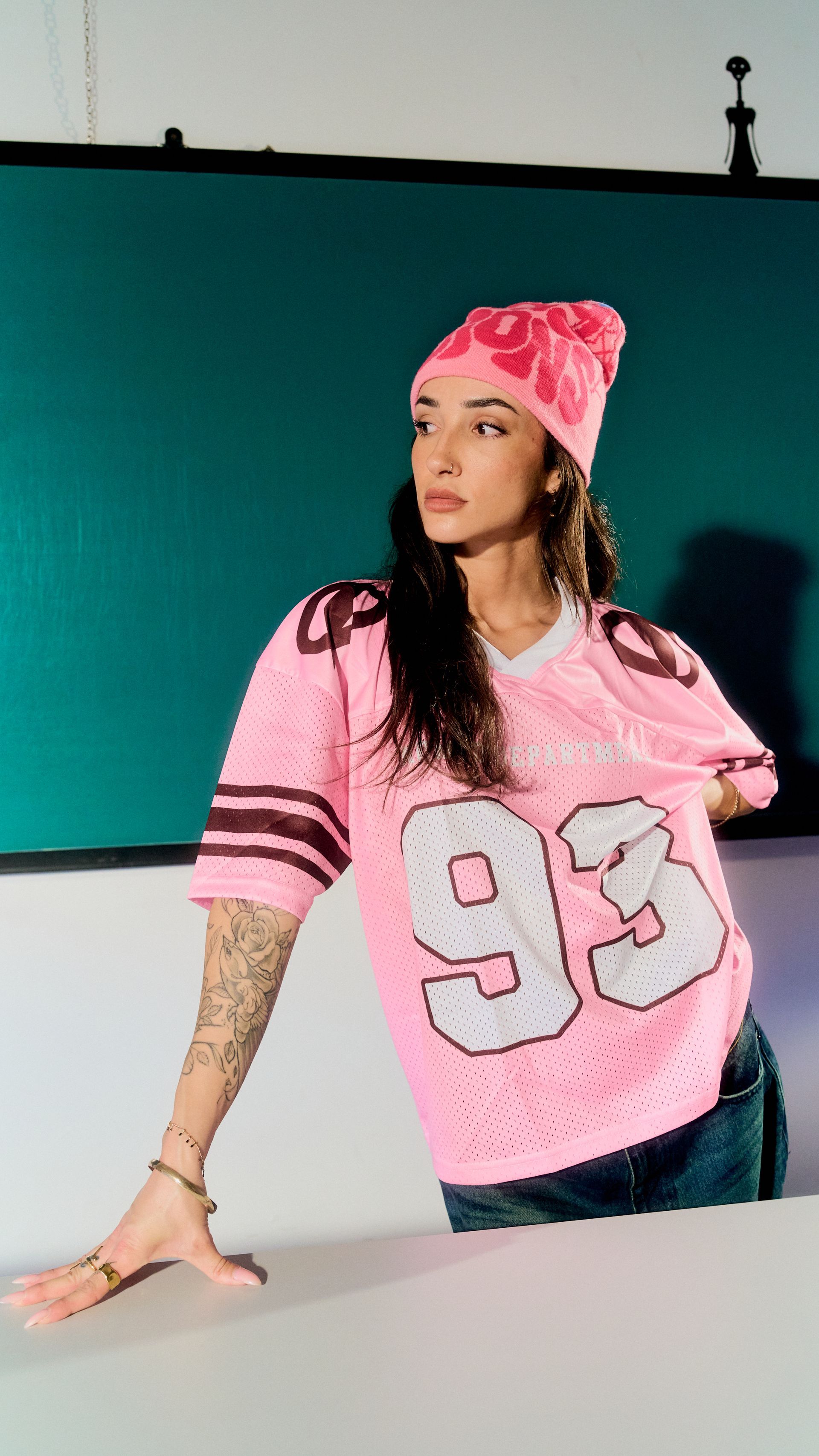 Mujer con jersey rosa y gorro de lana, apoyada en una mesa frente a un fondo azul verdoso.