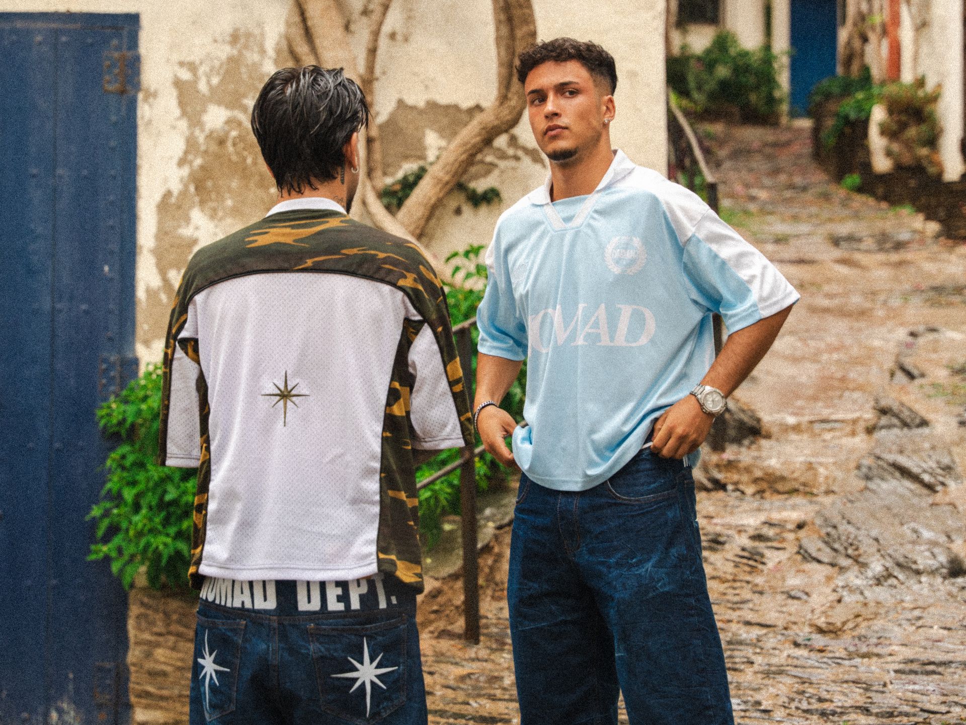 Dos hombres vestidos de manera informal conversan al aire libre en una calle adoquinada.