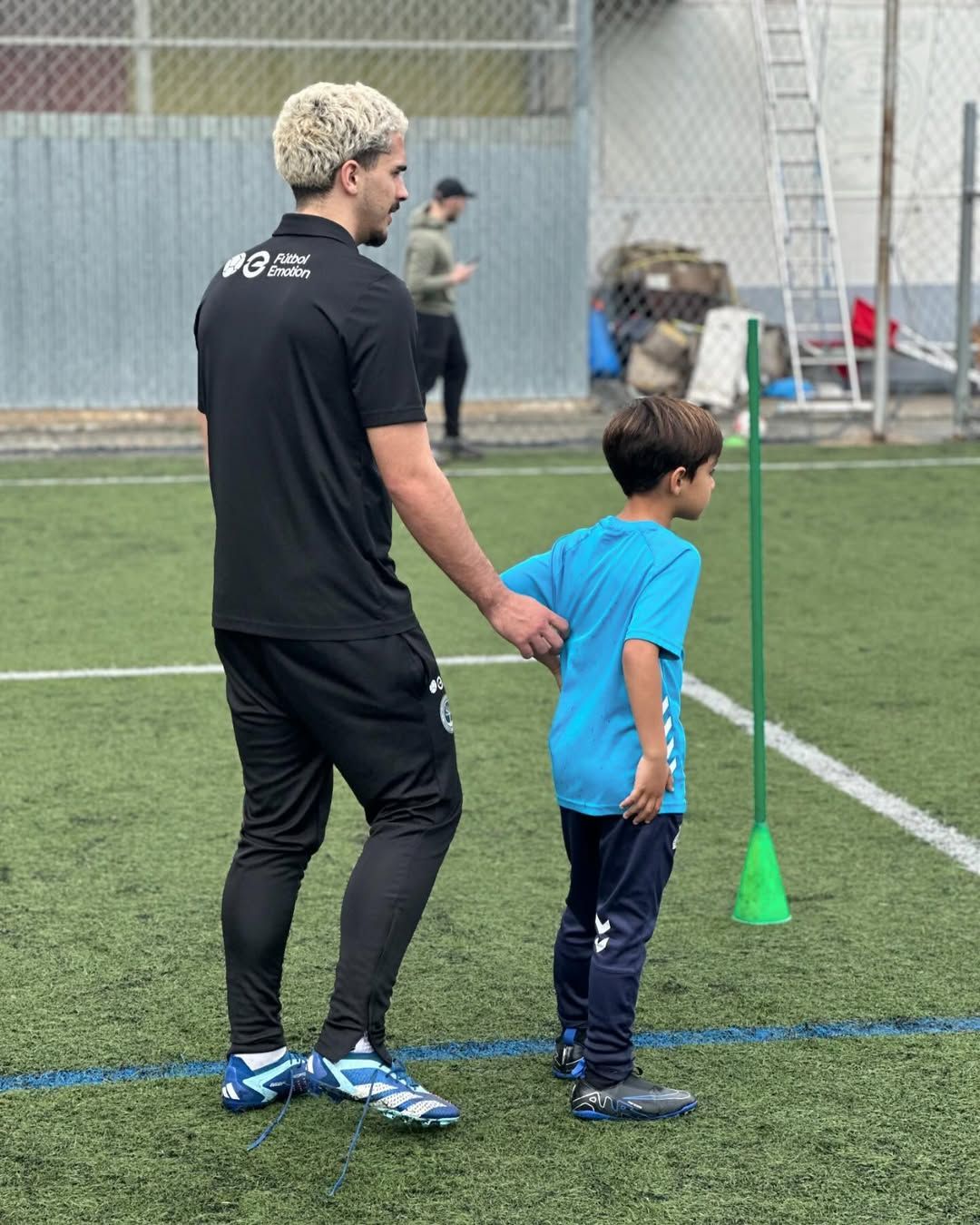 Un hombre de negro ayuda a un niño en un campo de fútbol. Están de pie cerca de un cono verde.
