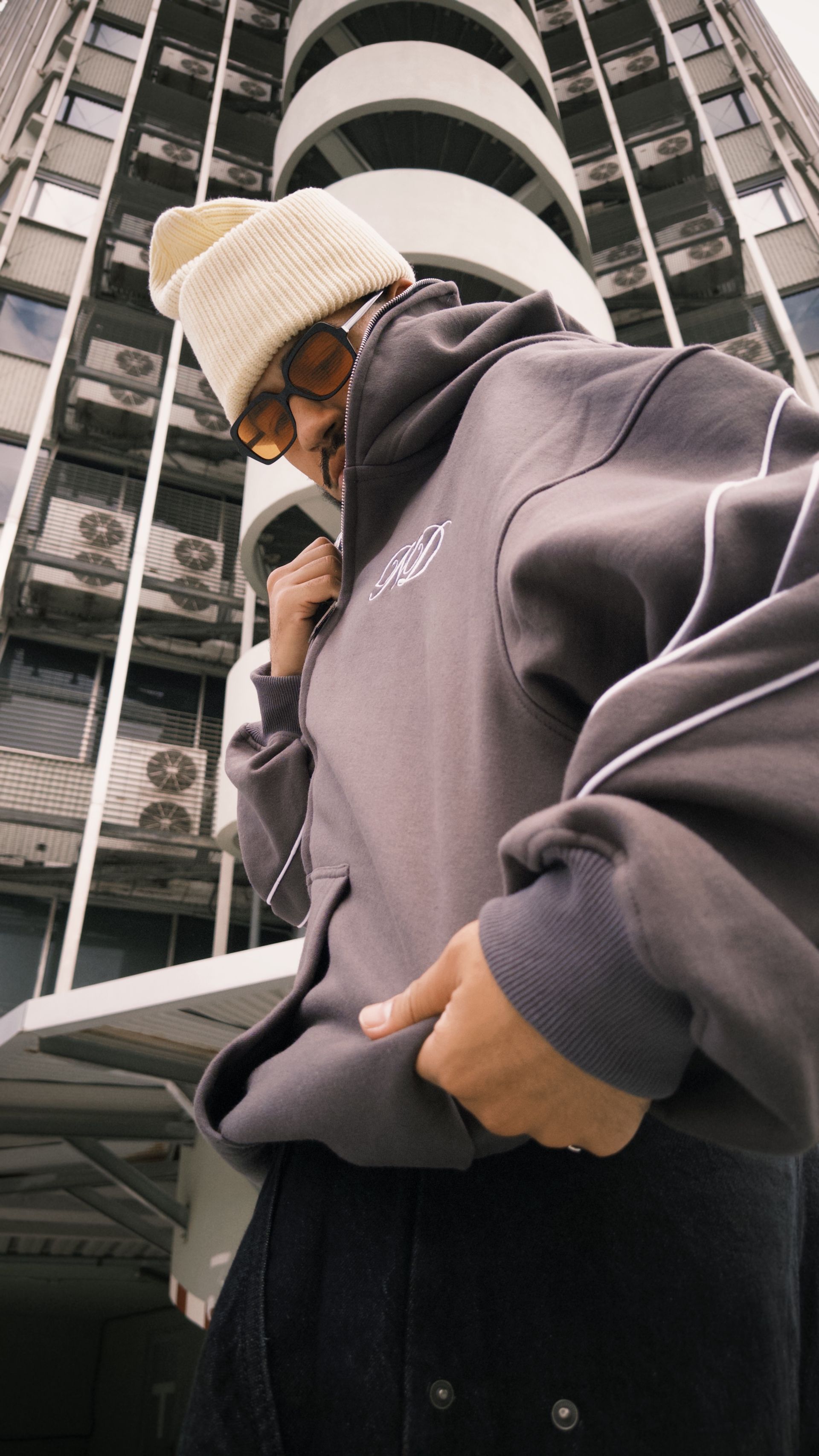 Persona con gorro y sudadera color beige señala hacia la cámara; al fondo se ve un edificio alto.