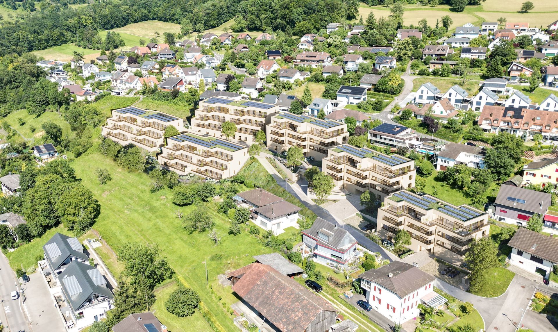 Luftaufnahme einer modernen Wohnanlage, umgeben von grünen Hügeln, Bäumen und kleinen Häusern in einer ländlichen Landschaft.