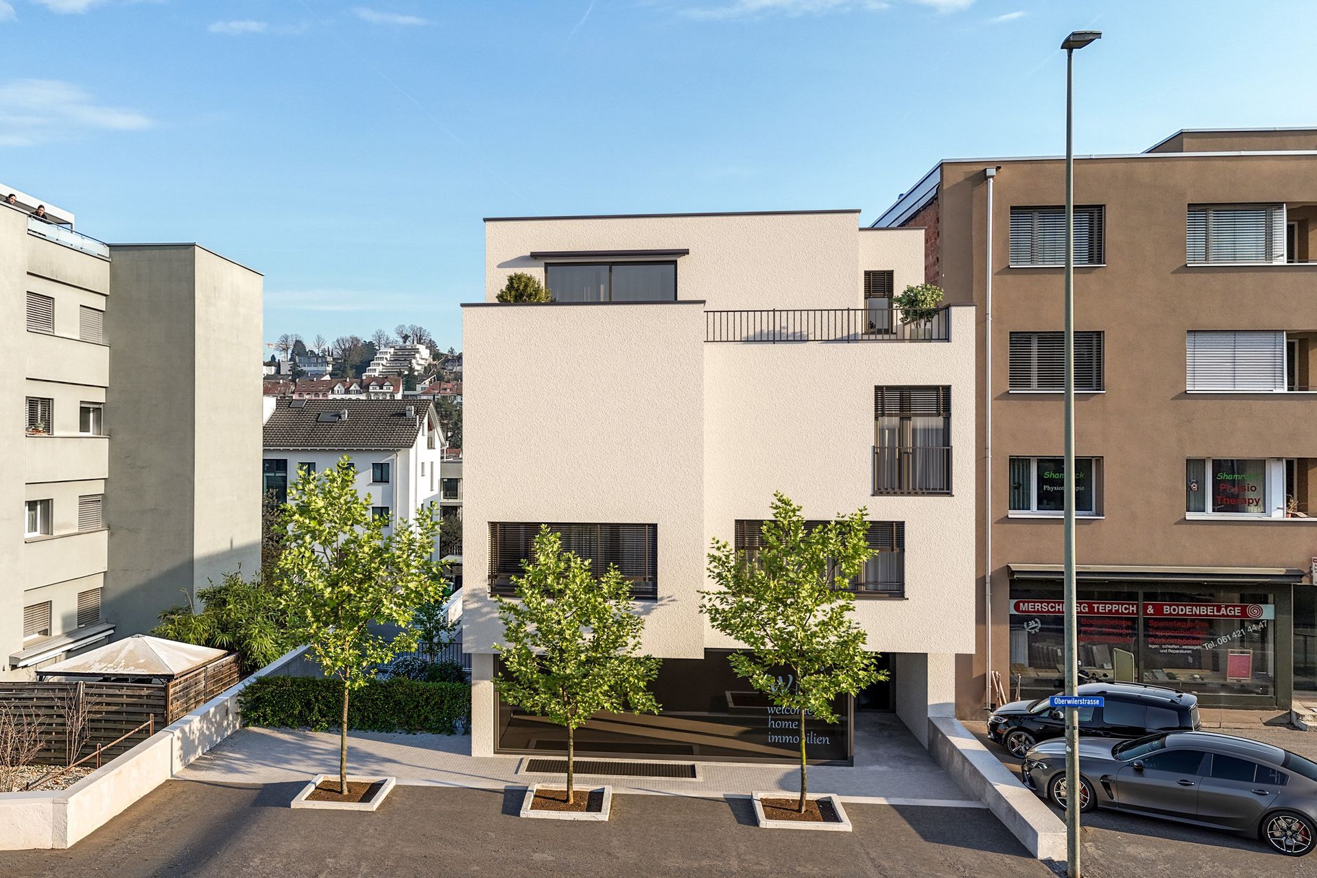 Zwei moderne, hellfarbige Wohnhäuser an einem sanften Hang, umgeben von Bäumen und einem gepflasterten Gehweg.