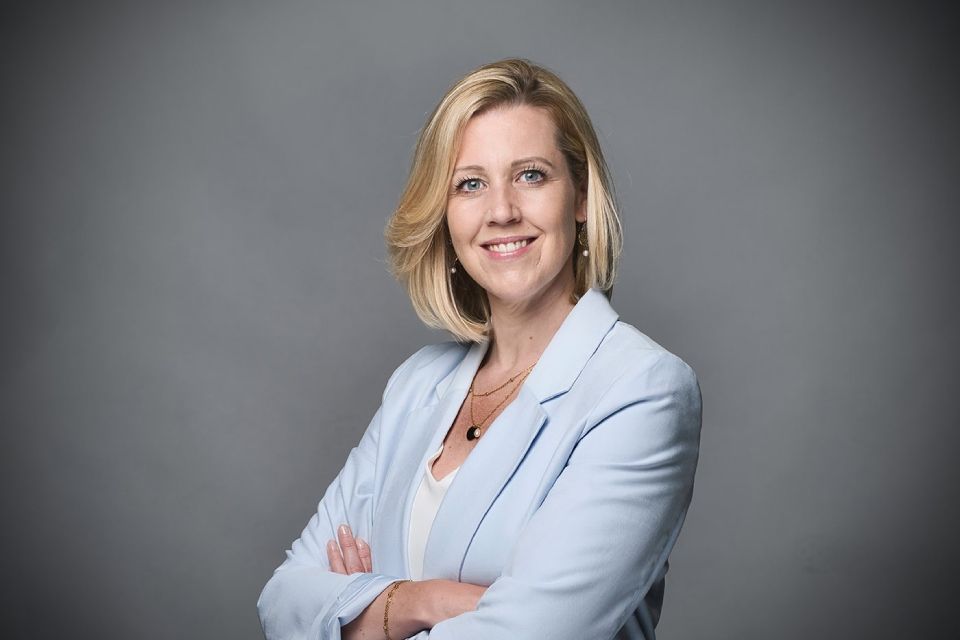 Eine professionelle Frau mit blonden Haaren, die einen hellblauen Blazer über einem weissen Top trägt, posiert mit verschränkten Armen.