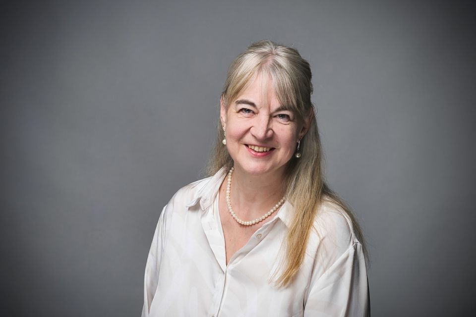 Eine Person mit langen blonden Haaren und einem weissen Hemd mit Kragen lächelt vor einem grauen Hintergrund.