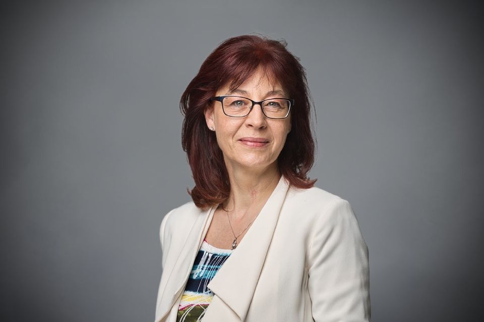 Eine Person mit kastanienbraunem Haar, Brille und hellem Blazer, blickt vor grauem Hintergrund in die Kamera.