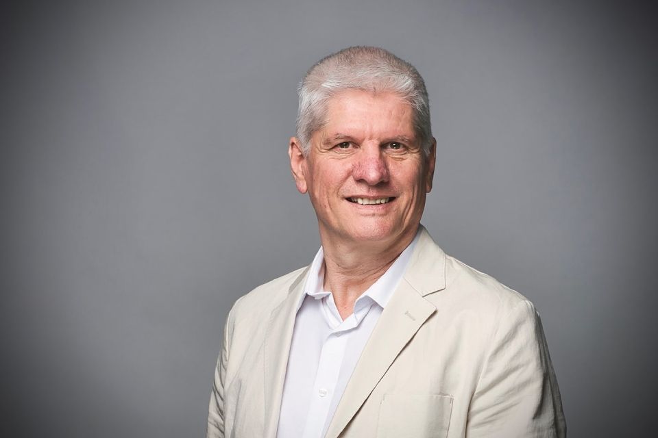 Ein Porträtfoto einer Person mit kurzen grauen Haaren, die ein weisses Hemd mit Kragen und einen beigen Blazer trägt, vor grauem Hintergrund.