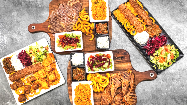 Trois planches en bois et une assiette garnie de viandes grillées, de riz jaune, de salades et de sauces, disposées sur une surface grise.